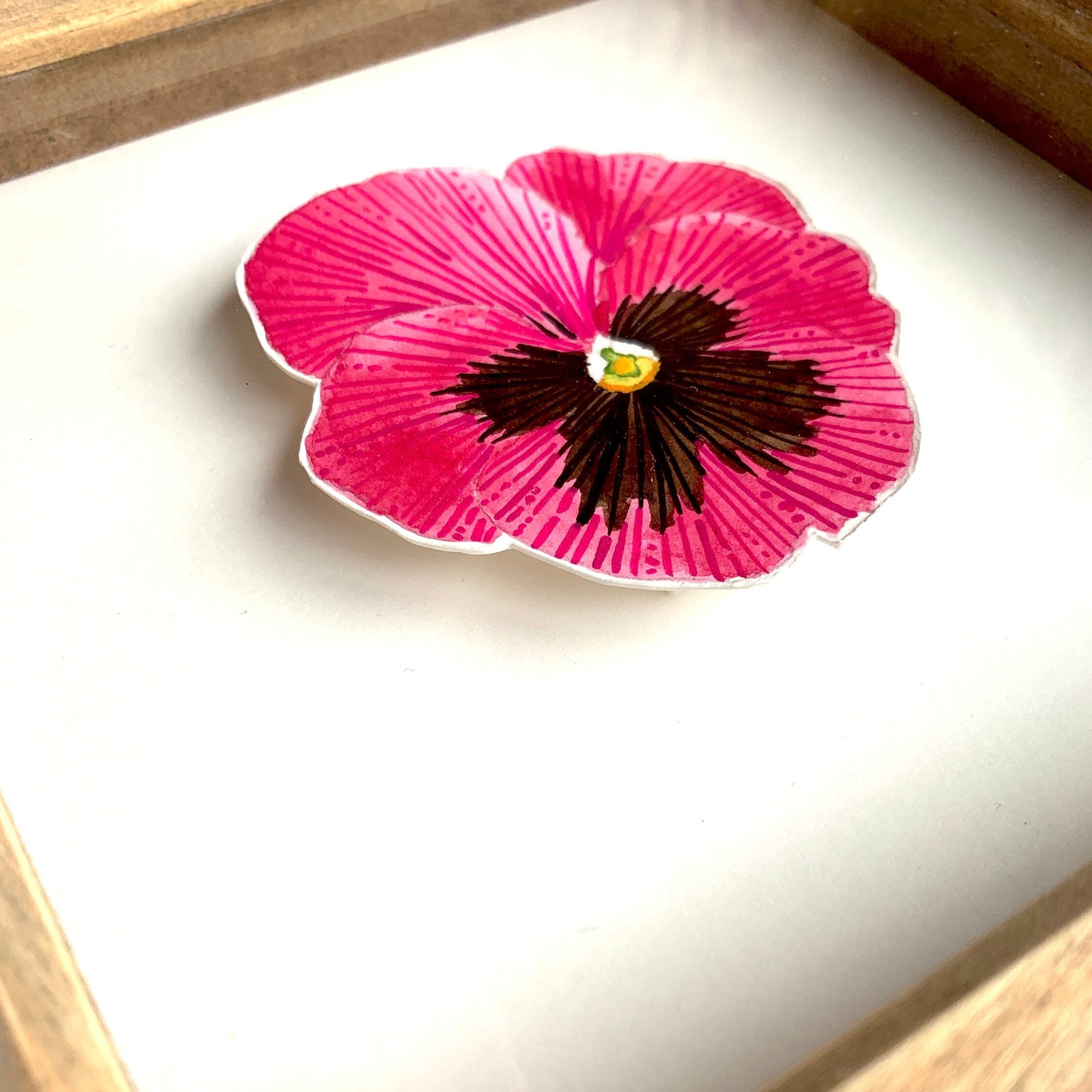 Pink pansy flower with a black center on a white card, framed in wood.
