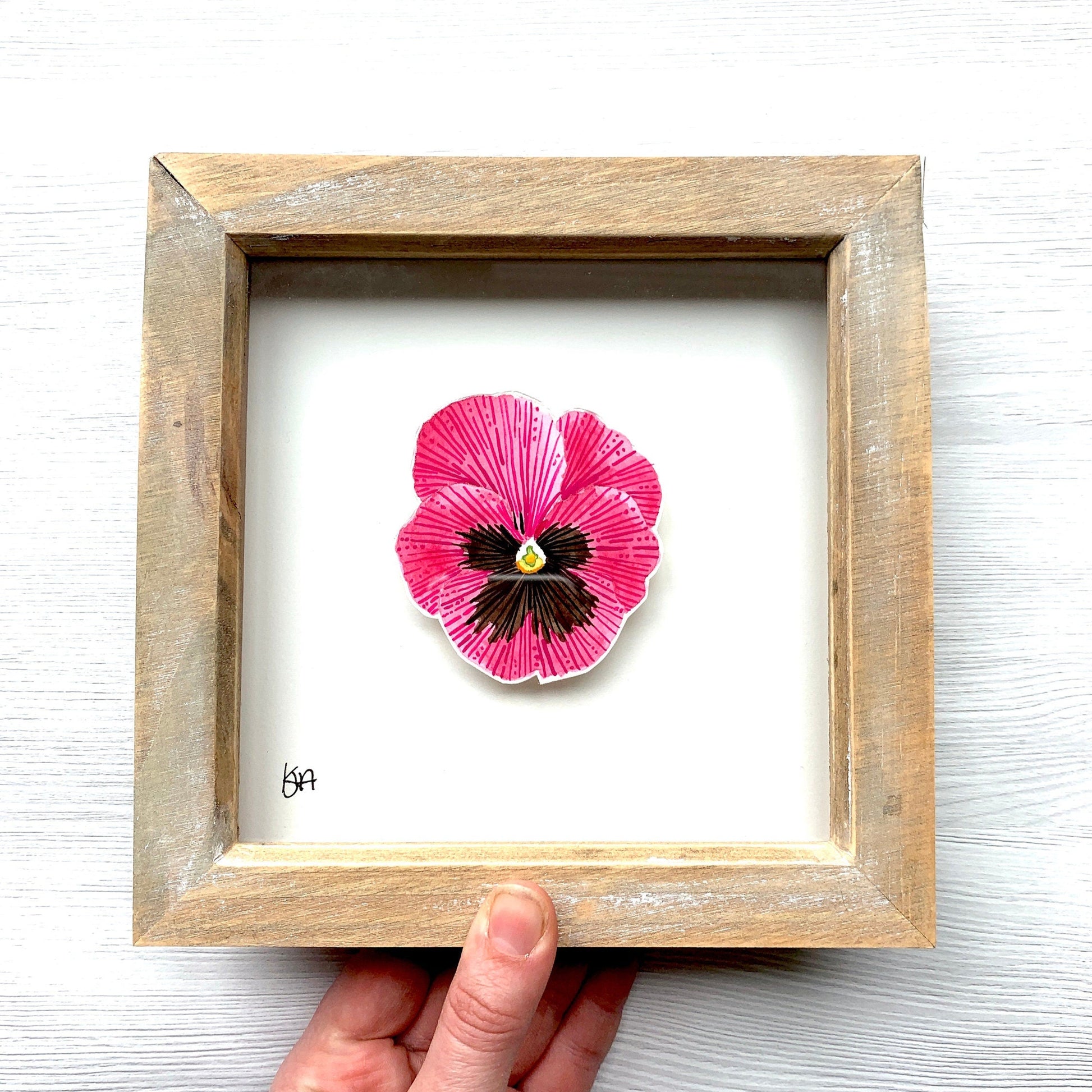 Framed artwork of a pink flower held by a hand on a white background