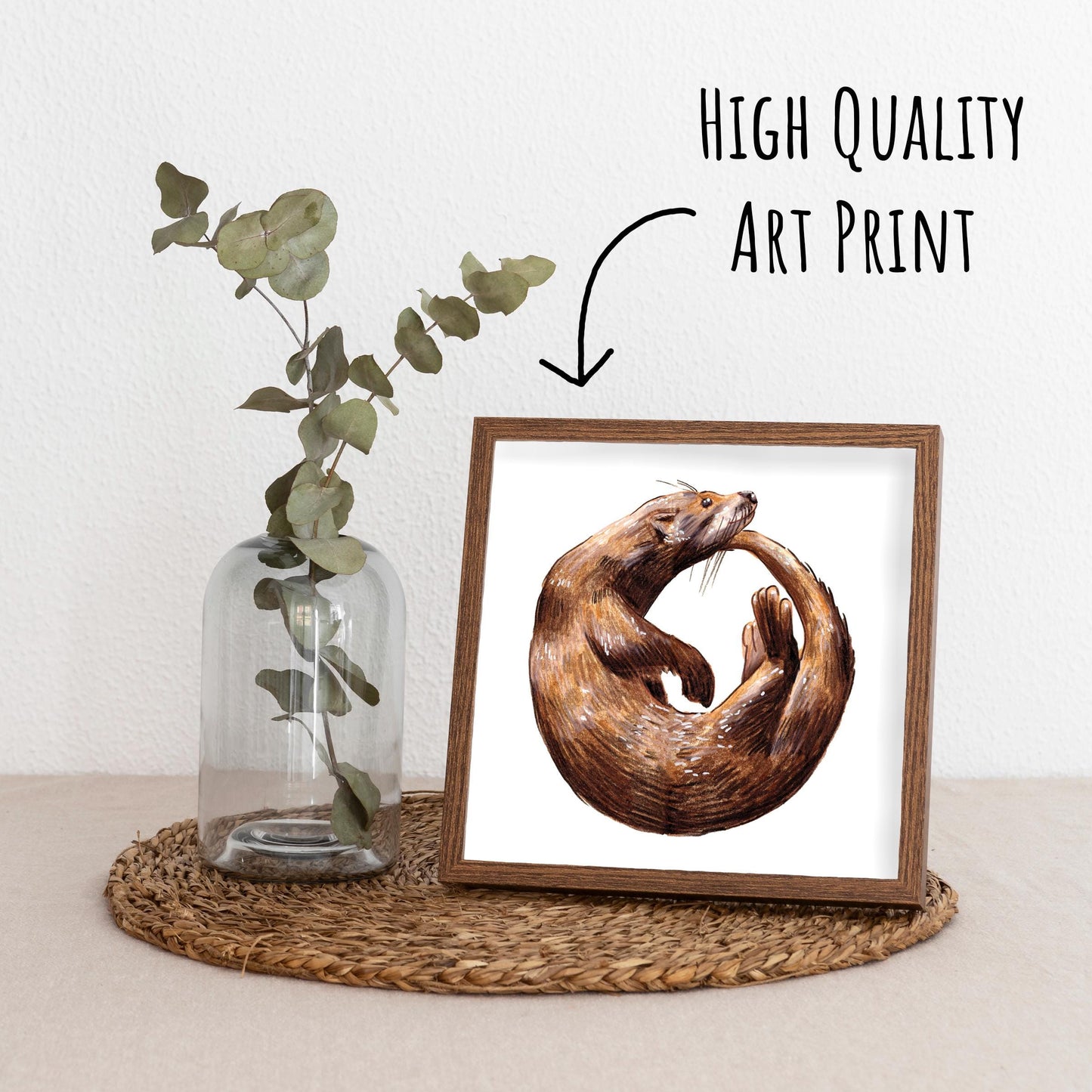 Playful Otter Art Print in a dark wood frame standing on a straw matt with a glass vase with a eucalyptus branch.