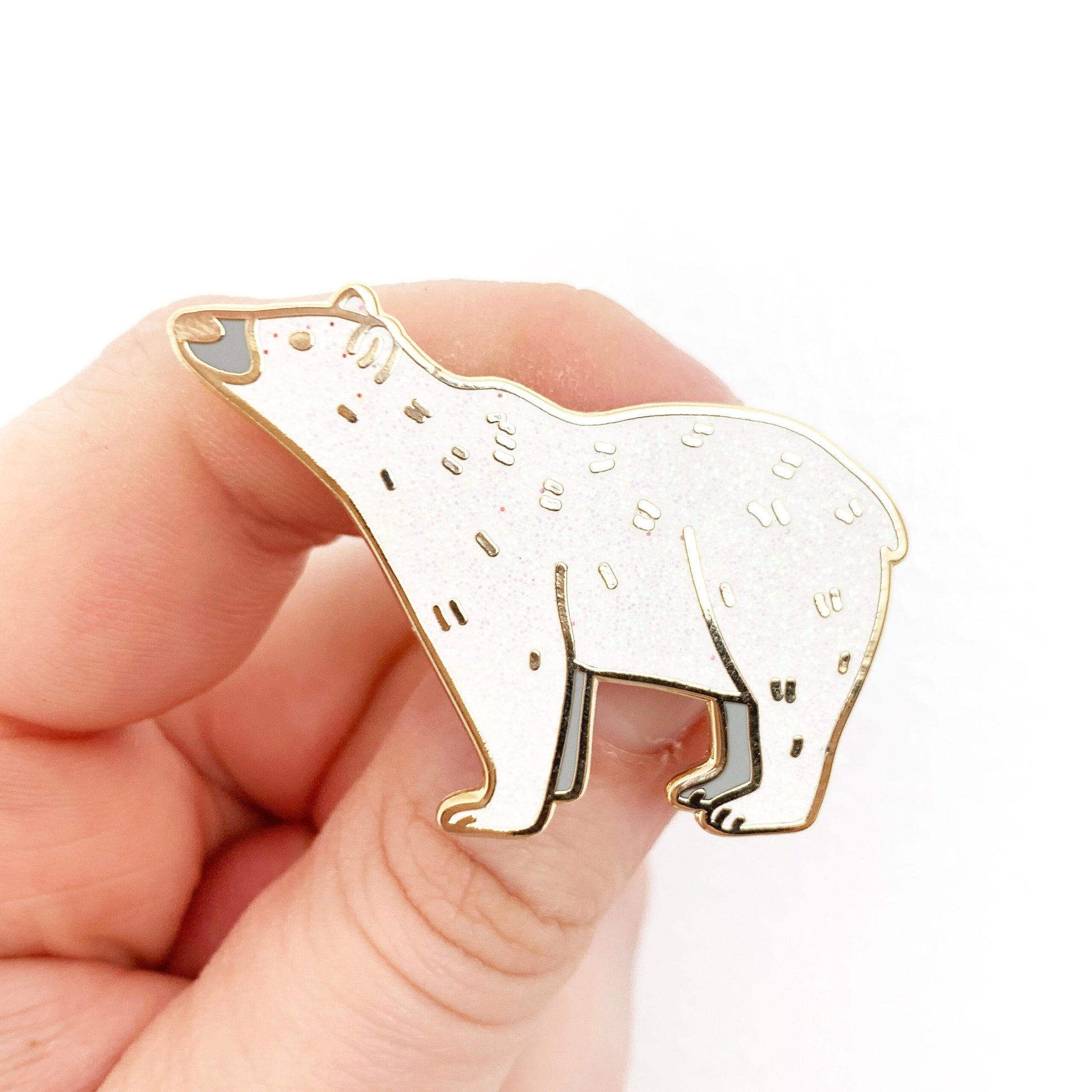 A gold-plated, grey and white glitter enamel polar bear pin badge held up against a white background.