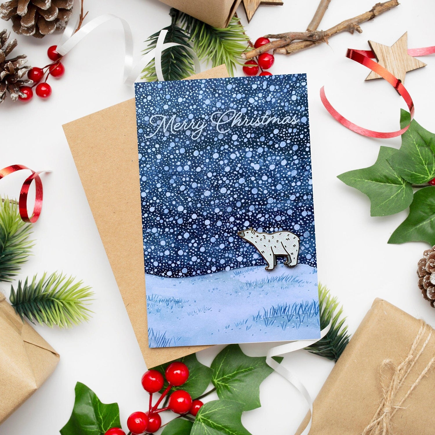 A Christmas card with a hand painted winter snowy scene featuring a polar bear, accompanied by a polar bear pin badge with white glitter and gold plating. The card is placed on a surface with festive decorations including red berries, green leaves, and brown kraft paper.