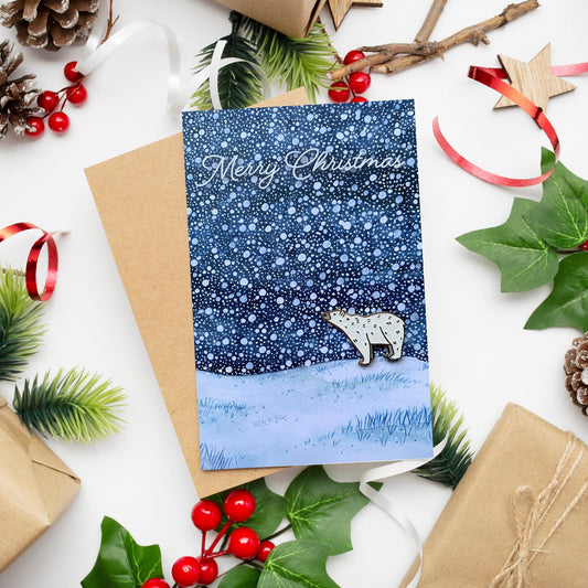 A Christmas card with a hand painted winter snowy scene featuring a polar bear, accompanied by a polar bear pin badge with white glitter and gold plating. The card is placed on a surface with festive decorations including red berries, green leaves, and brown kraft paper.