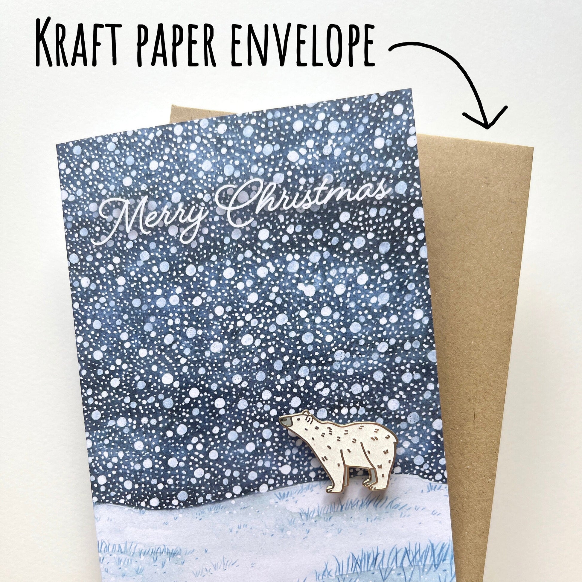 Card with 'Merry Christmas' text and a glittery polar bear pin badge, held over a kraft paper envelope on a white background.