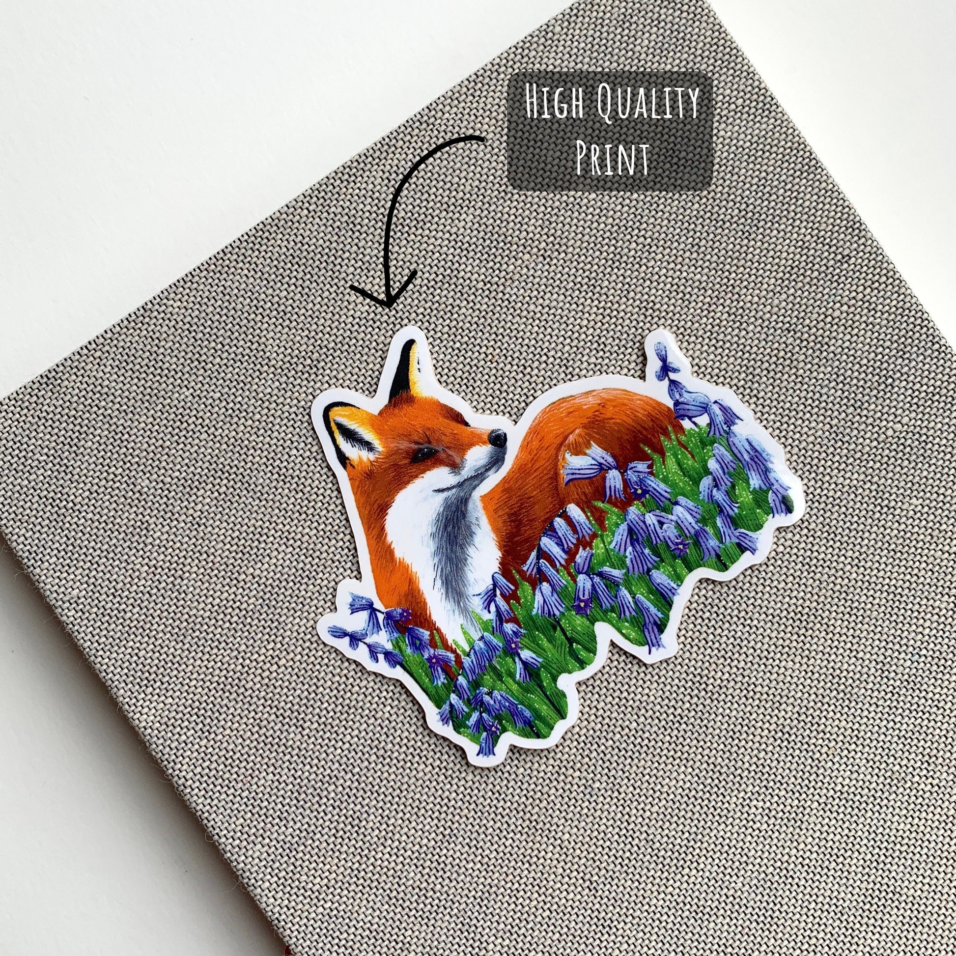 Red Fox and Bluebells Sticker placed on a grey notebook with the text high quality print.