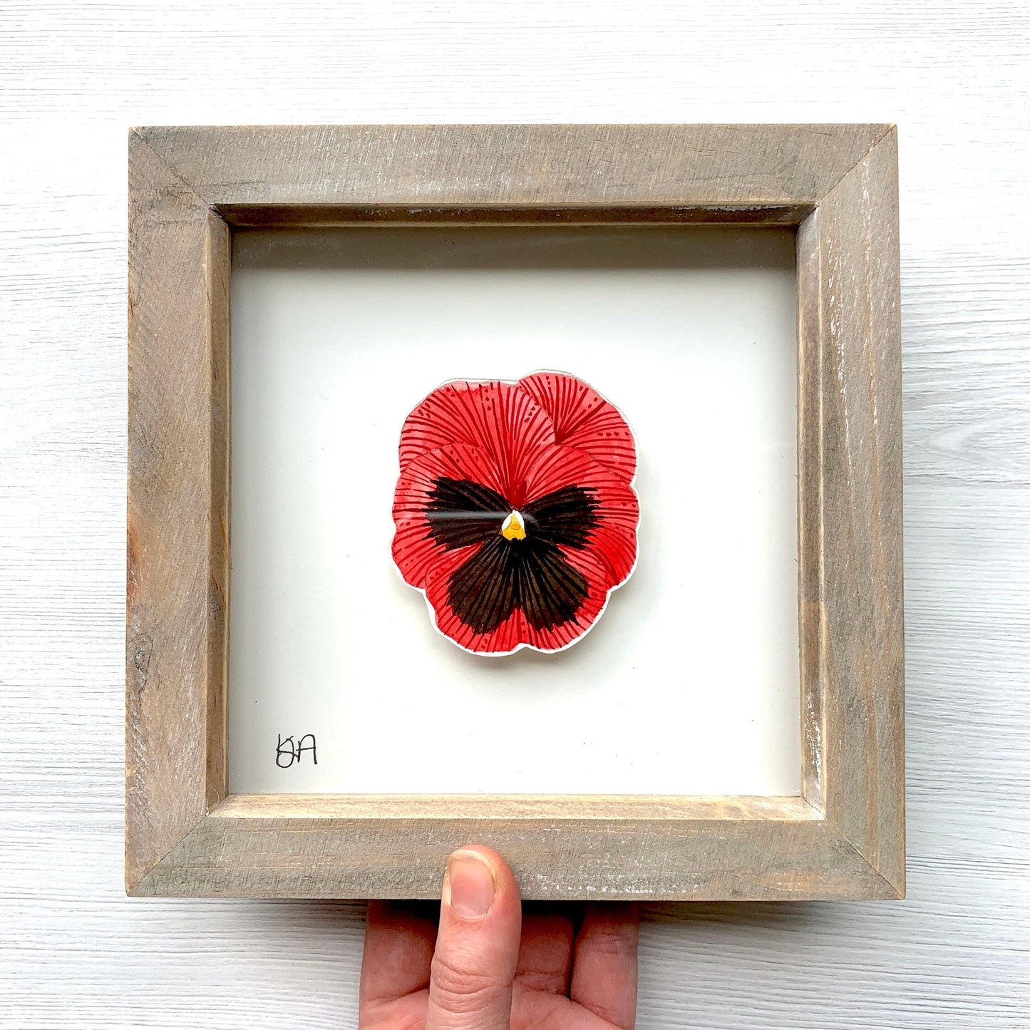 Framed artwork of a red and black flower held by a hand on a white background