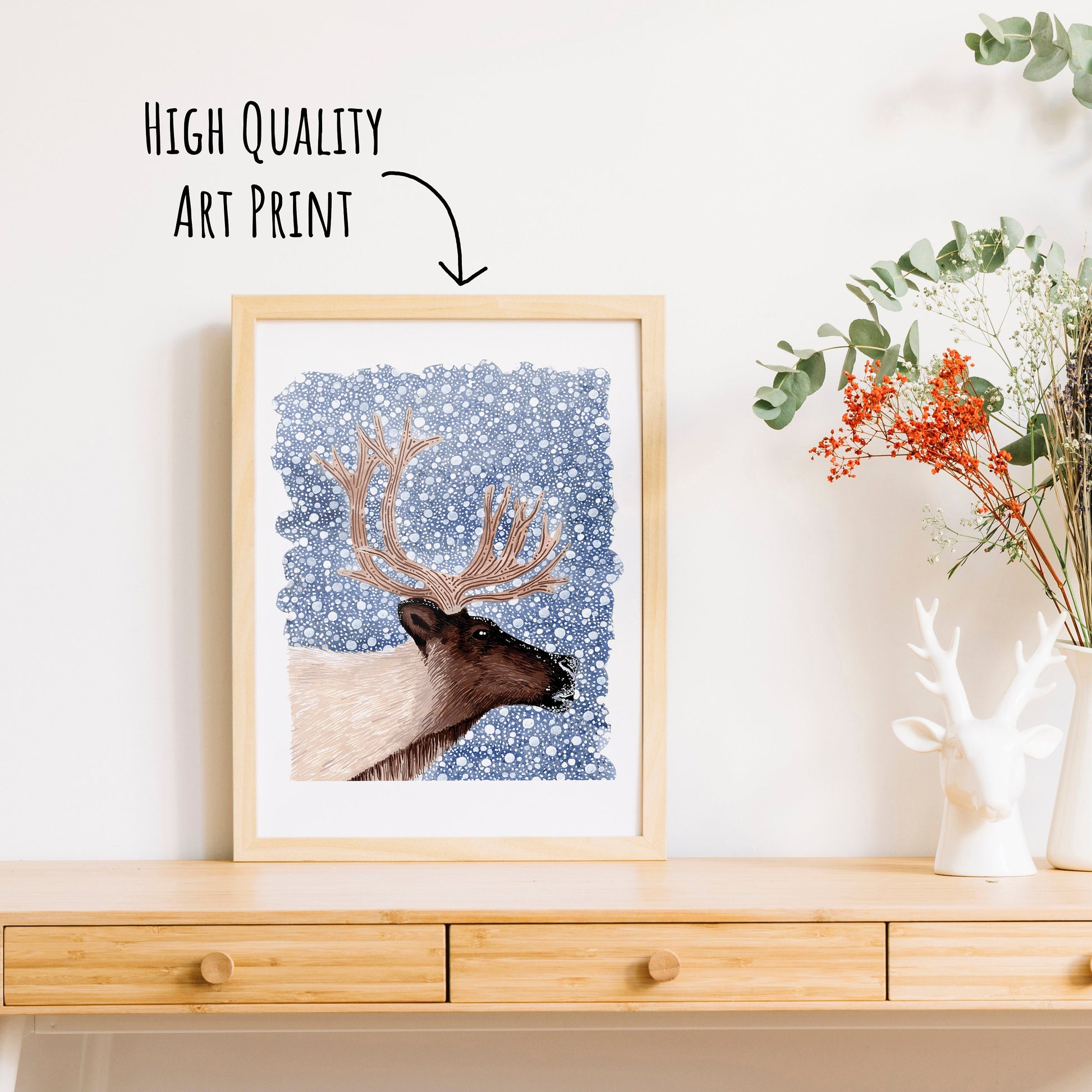A reindeer art print with a dark blue snowy background in a frame. On a table top with a vase of dried foliage.