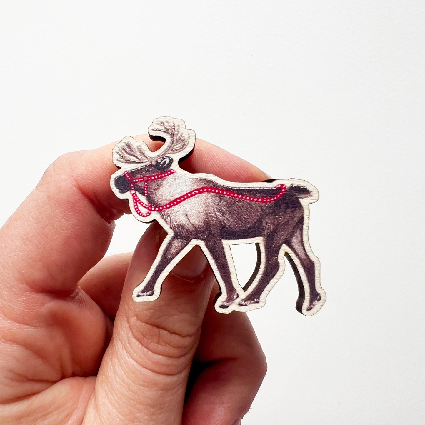 A hand holding a reindeer wooden pin badge design, A brown reindeer with bright red reins illustration. 