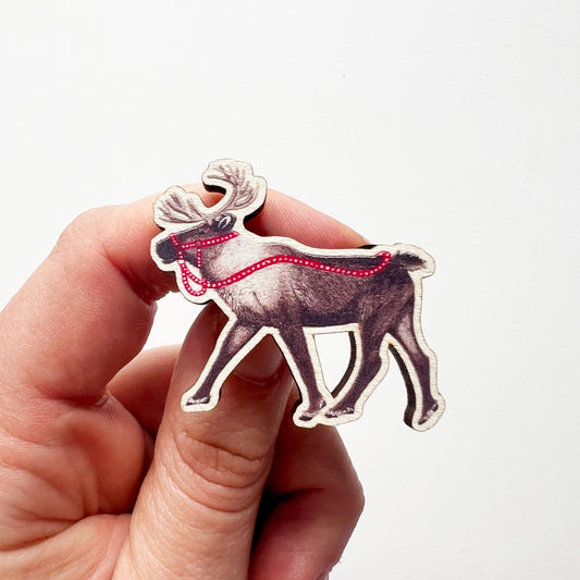 A hand holding a reindeer wooden pin badge design, A brown reindeer with bright red reins illustration. 