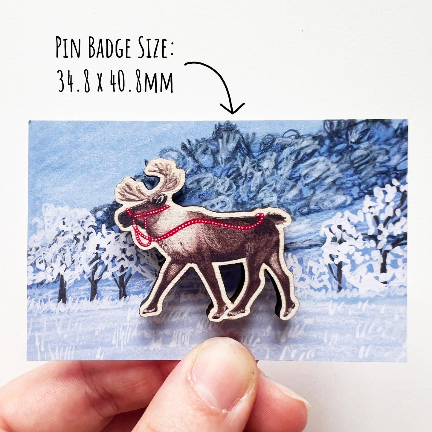 Reindeer Maple Wood Pin Badge shown on its frost winter landscape scene backing backing. being held up against a white background with the text pin badge size: 34.8 x 40.80 mm.
