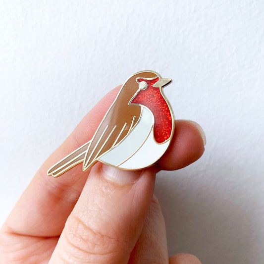 A hand holding a robin enamel pin badge with gold plating and red glitter enamel detailing. Held up against a white background.