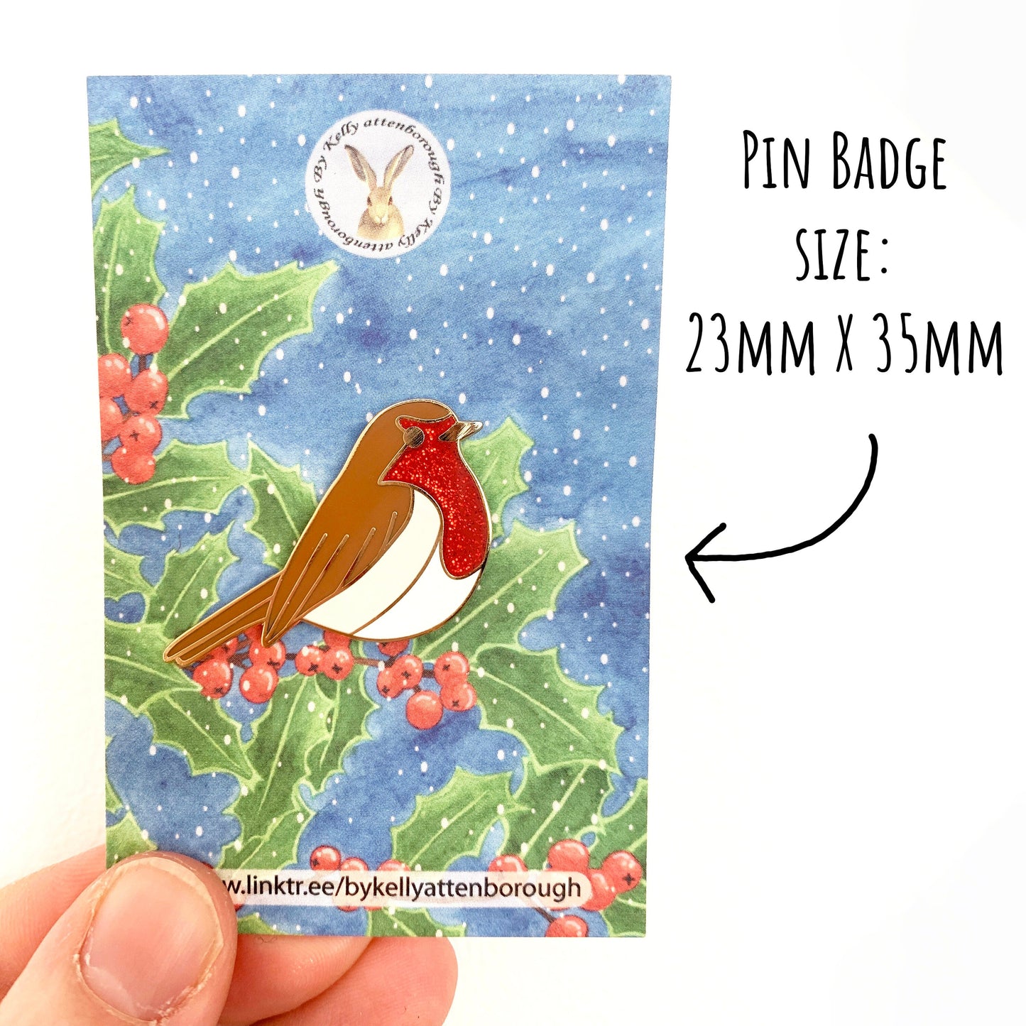 Robin Enamel Pin Badge on a backing card illustrated with holly. being held up against a white background with the text pin badge size: 23mm x 35mm.