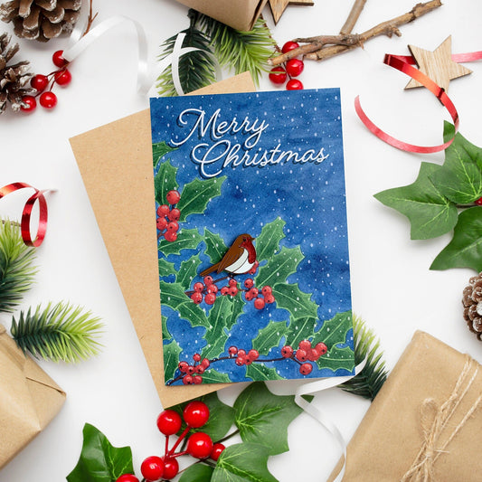 A Christmas card with a painting of a robin on holly leaves and berries, with snow and 'Merry Christmas' text, accompanied by a kraft paper envelope and festive decorations.