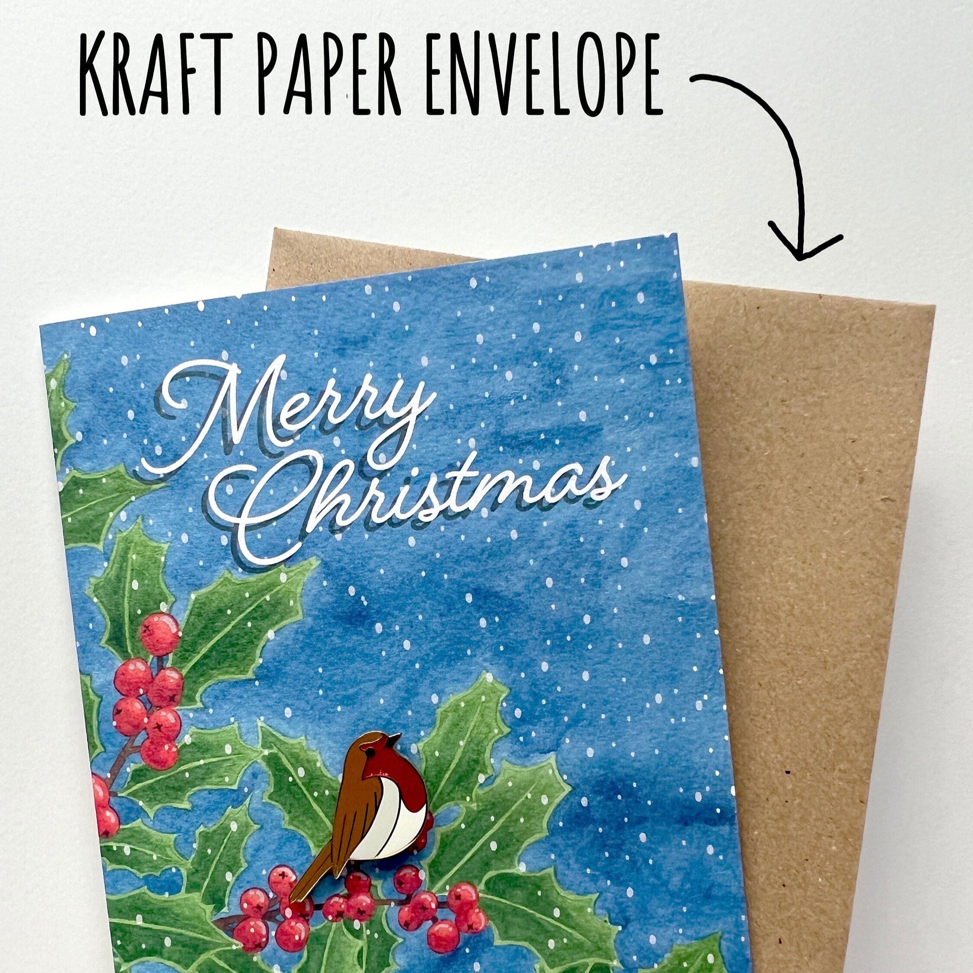 Christmas card with a robin pin badge and holly design on blue background, next to a kraft paper envelope.