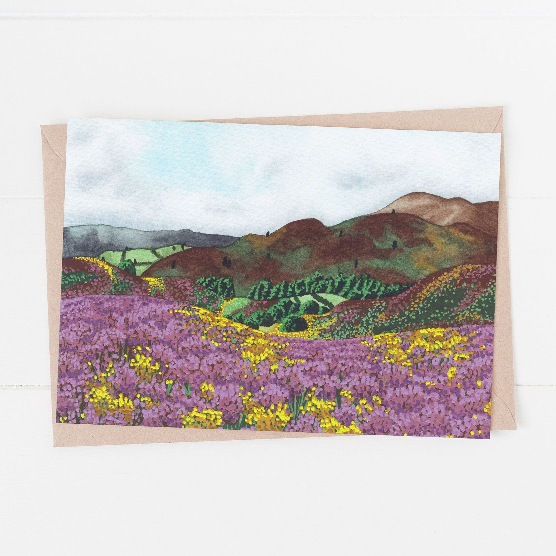 A greeting card featuring a vibrant illustration of a countryside landscape with fields of purple and yellow flowering heathers, hills, and a blue sky. The card has a beige border and the occasion is not specified.