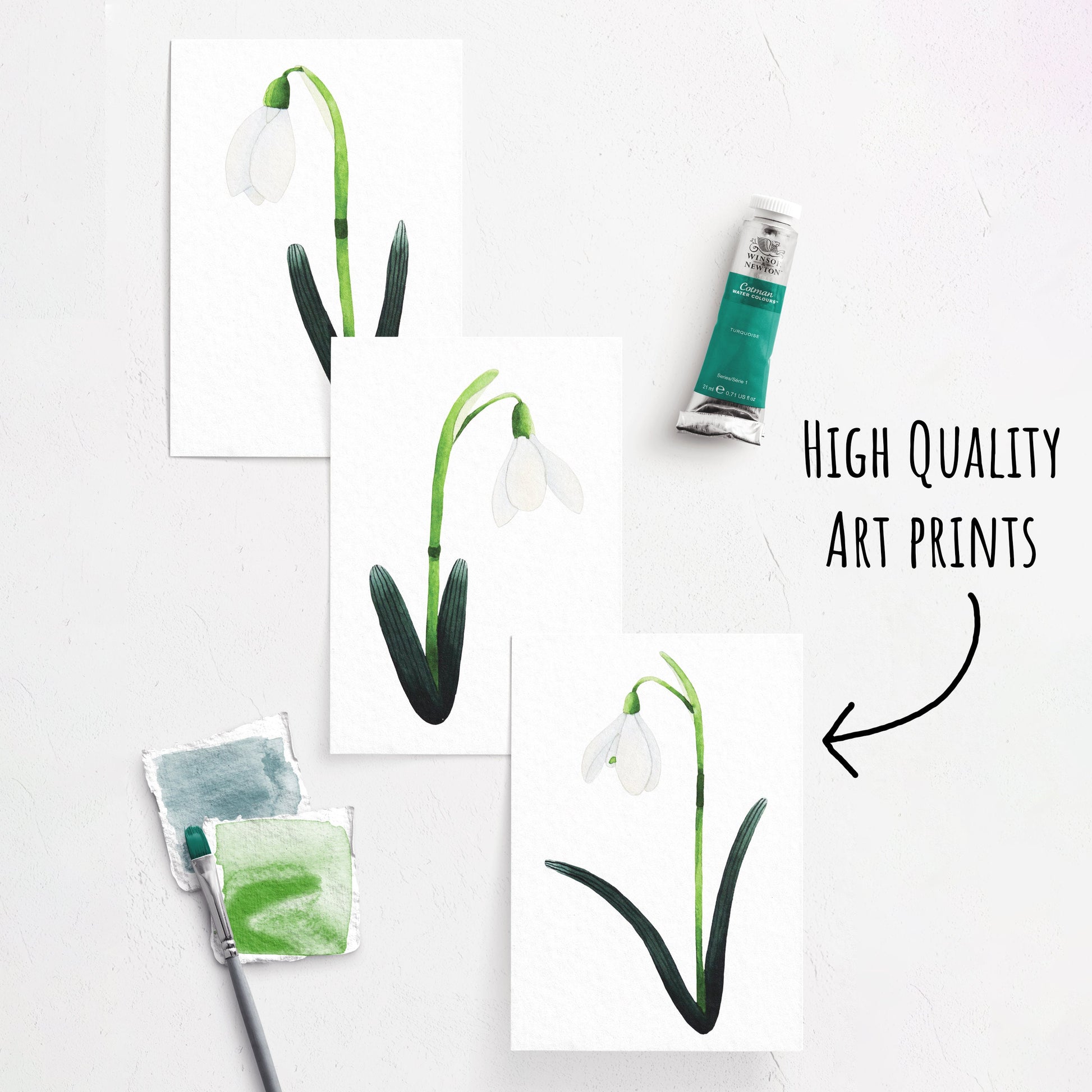 showing three of the Snowdrop Art Prints laying flat on a white surface. whit a paint brush and paint tube next to them.