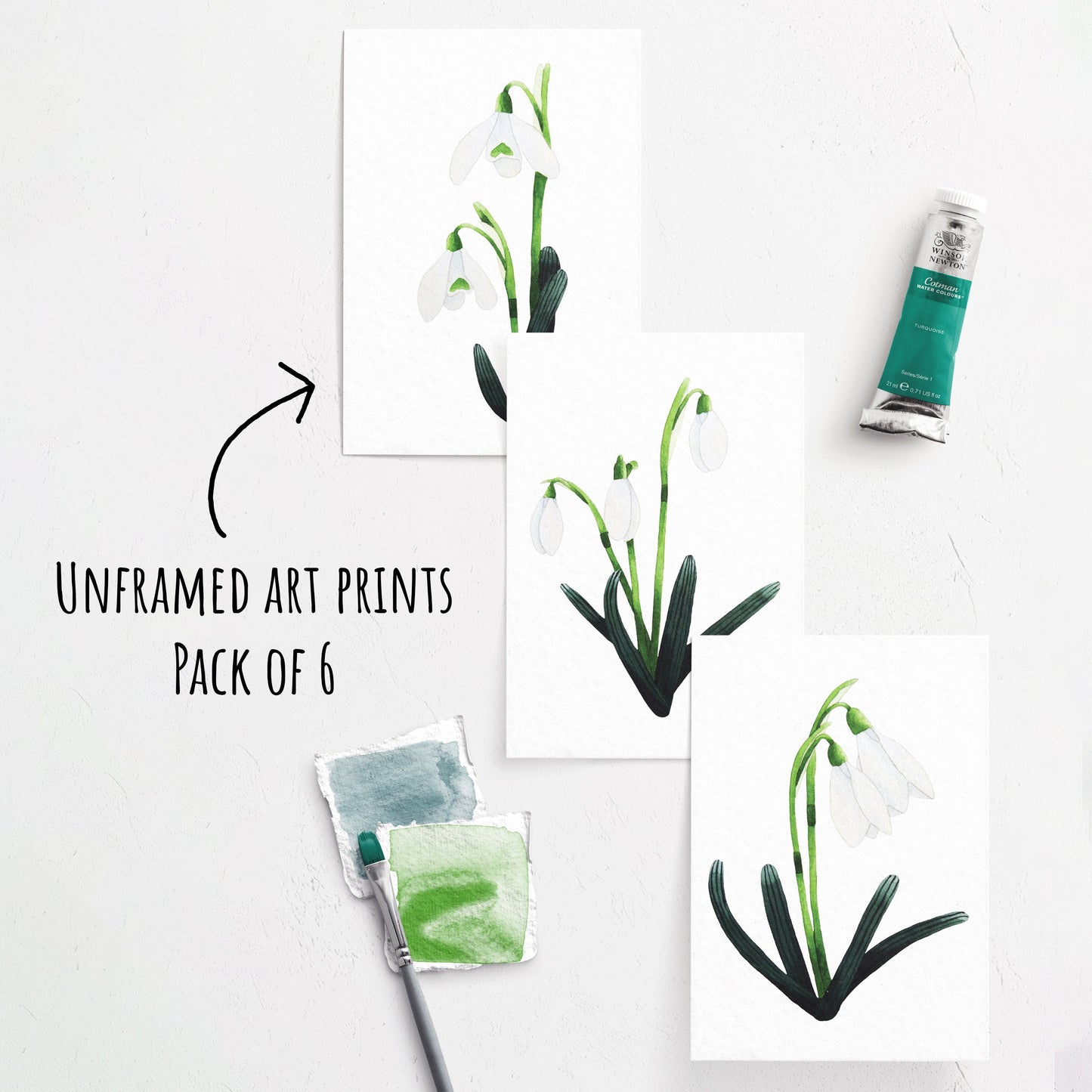 showing three of the Snowdrop Art Prints laying flat on a white surface. whit a paint brush and paint tube next to them.