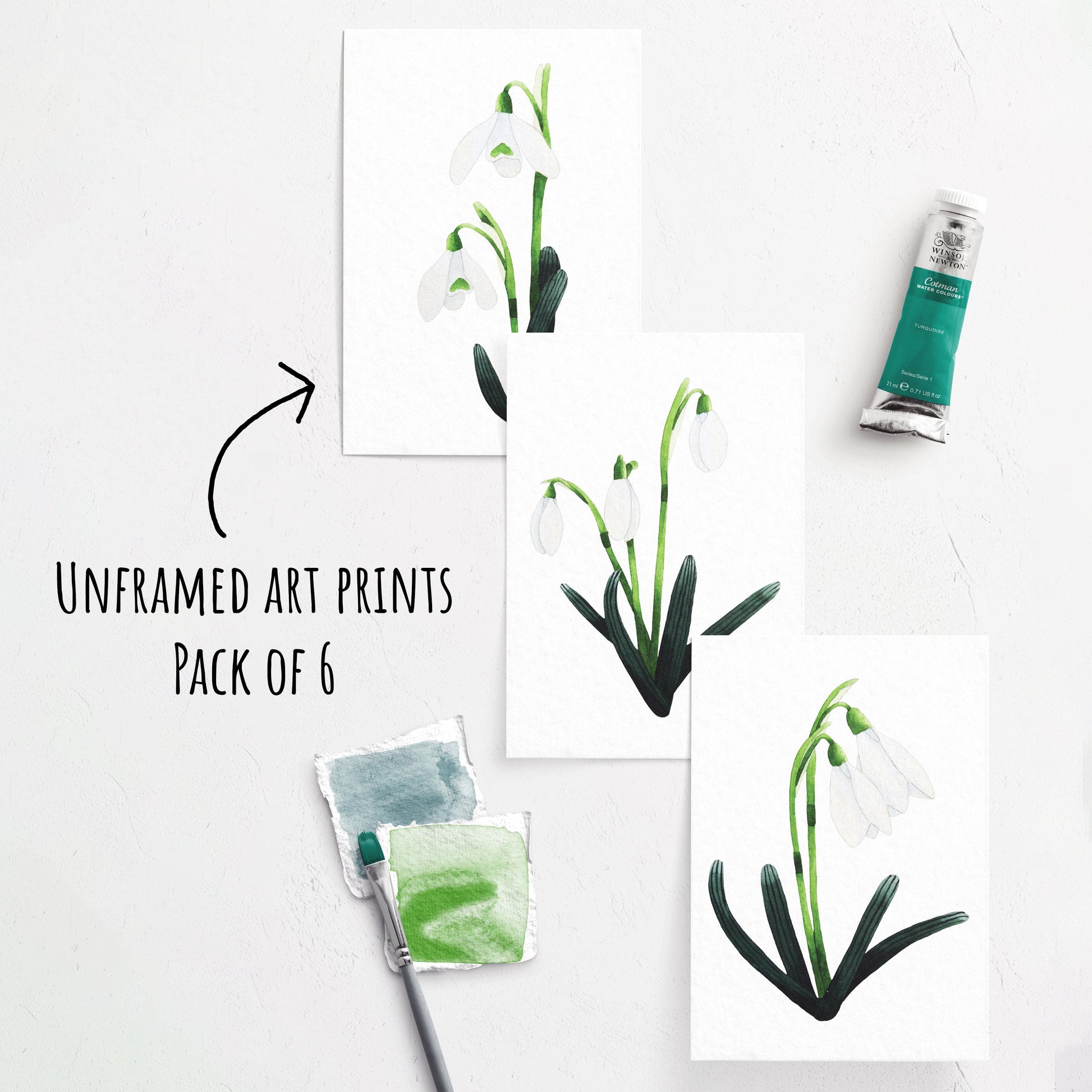 showing three of the Snowdrop Art Prints laying flat on a white surface. whit a paint brush and paint tube next to them.