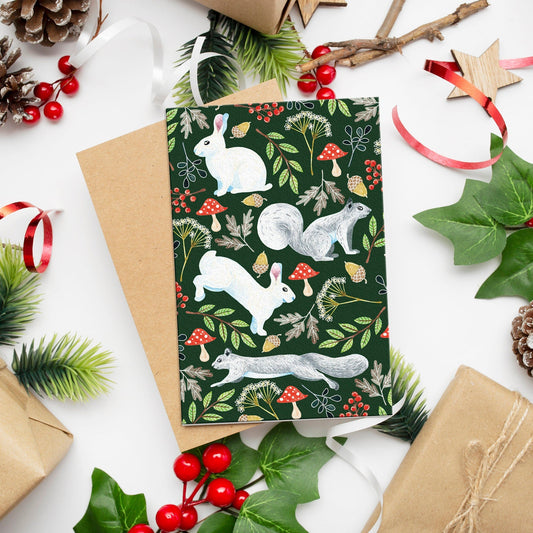Squirrel & Rabbit Christmas Card illustrated with rabbits, squirrels, woodland foliage with a kraft paper envelope On a white background surround by Christmas foliage.