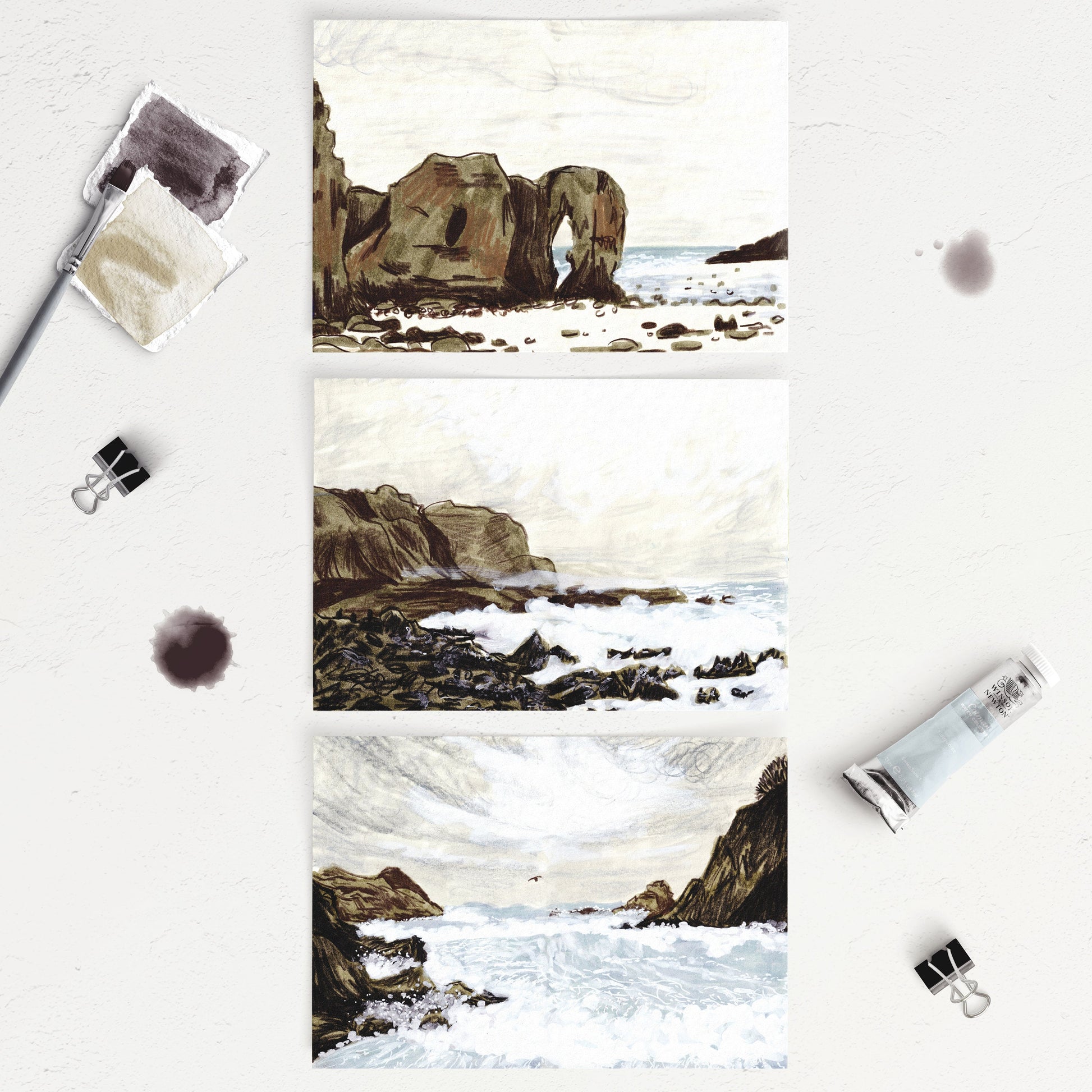A set of three A6 sized postcards featuring different stormy seascape illustrations with rock formations, placed on a table with painting supplies around them.
