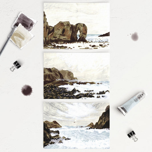 A set of three A6 sized postcards featuring different stormy seascape illustrations with rock formations, placed on a table with painting supplies around them.
