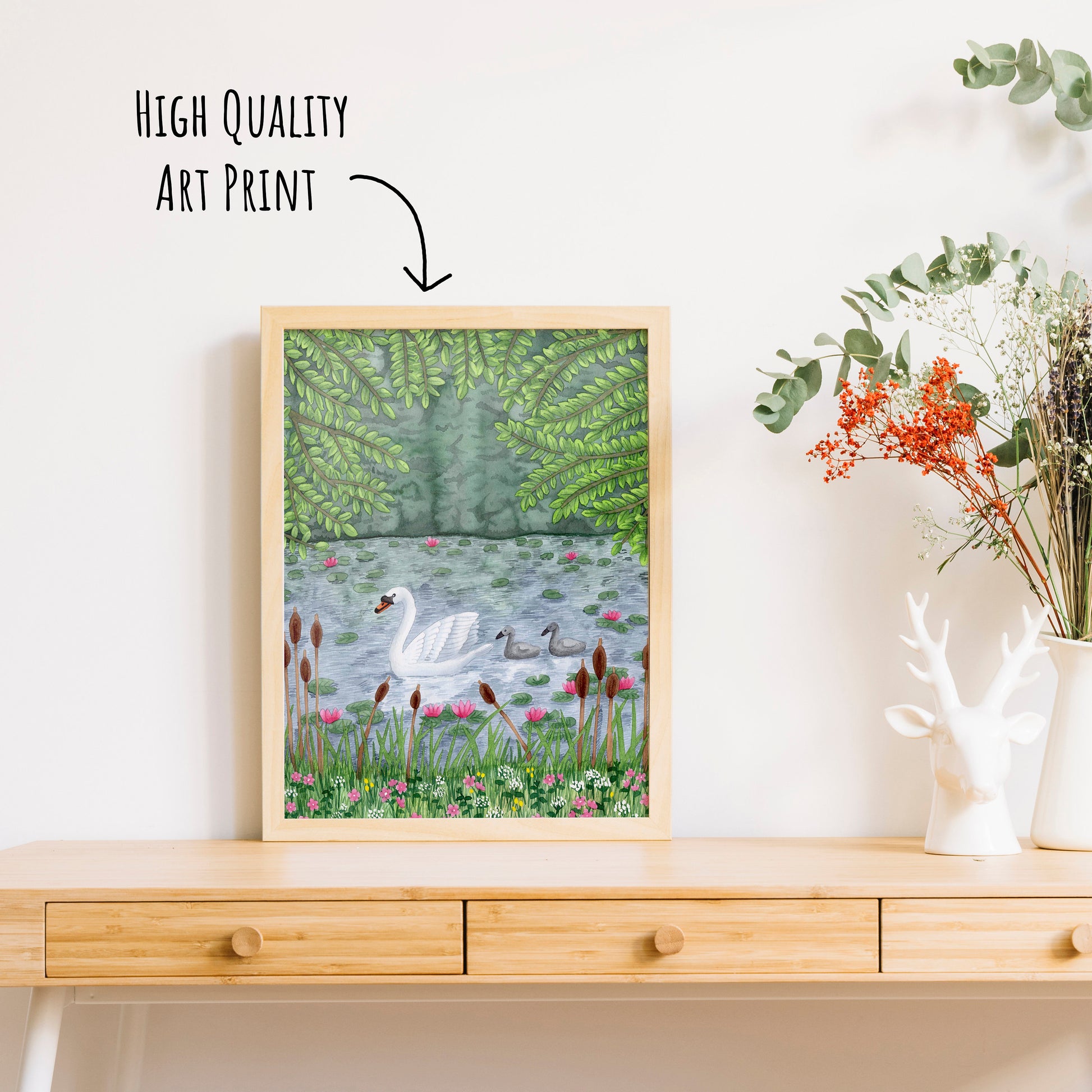 Swan and Cygnet Art Print shown in a frame on a table top with a vase for dried foliage.  (frame not included in purchase)