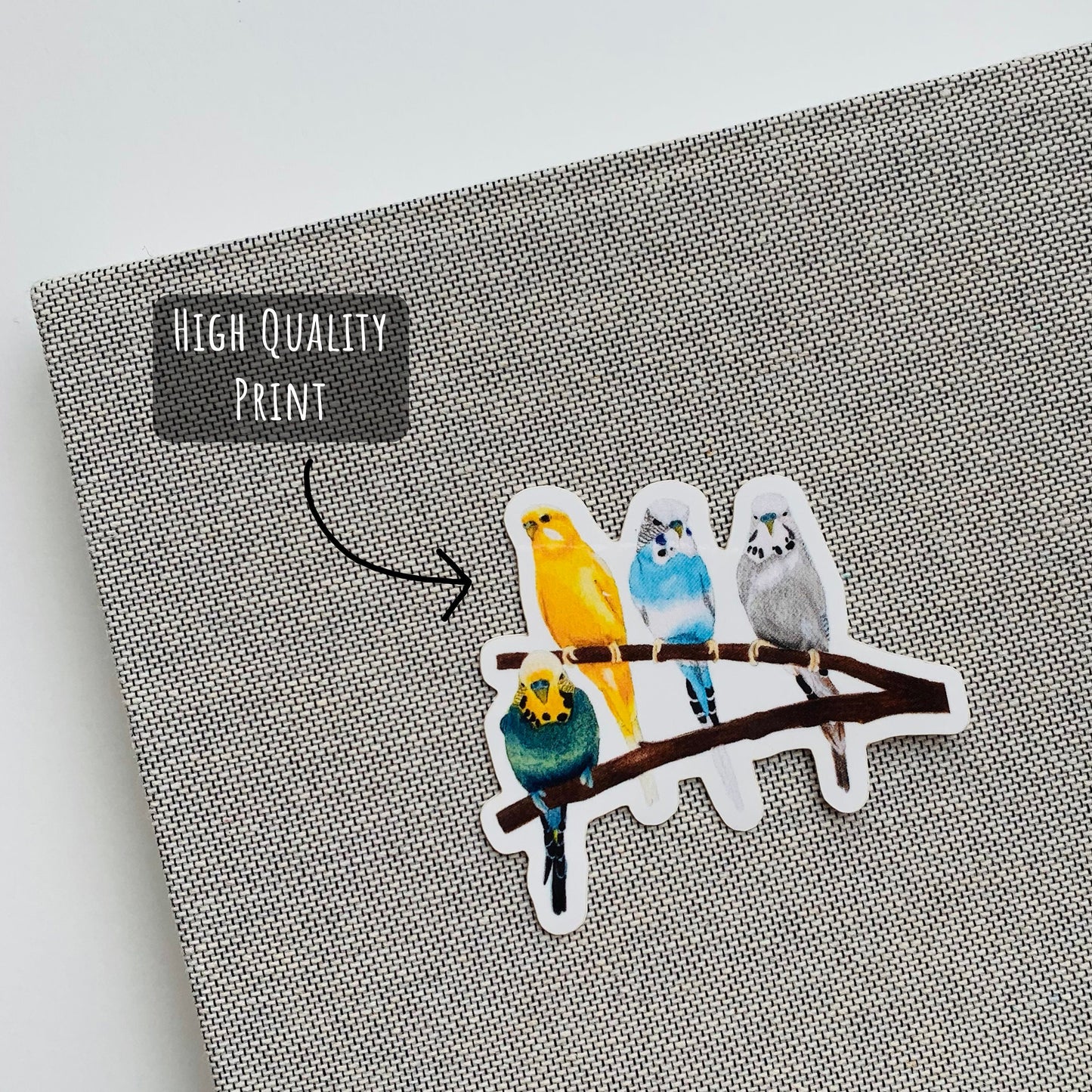 Vibrant Budgie Birds sticker placed on the front of a grey notebook with the text high quality print.