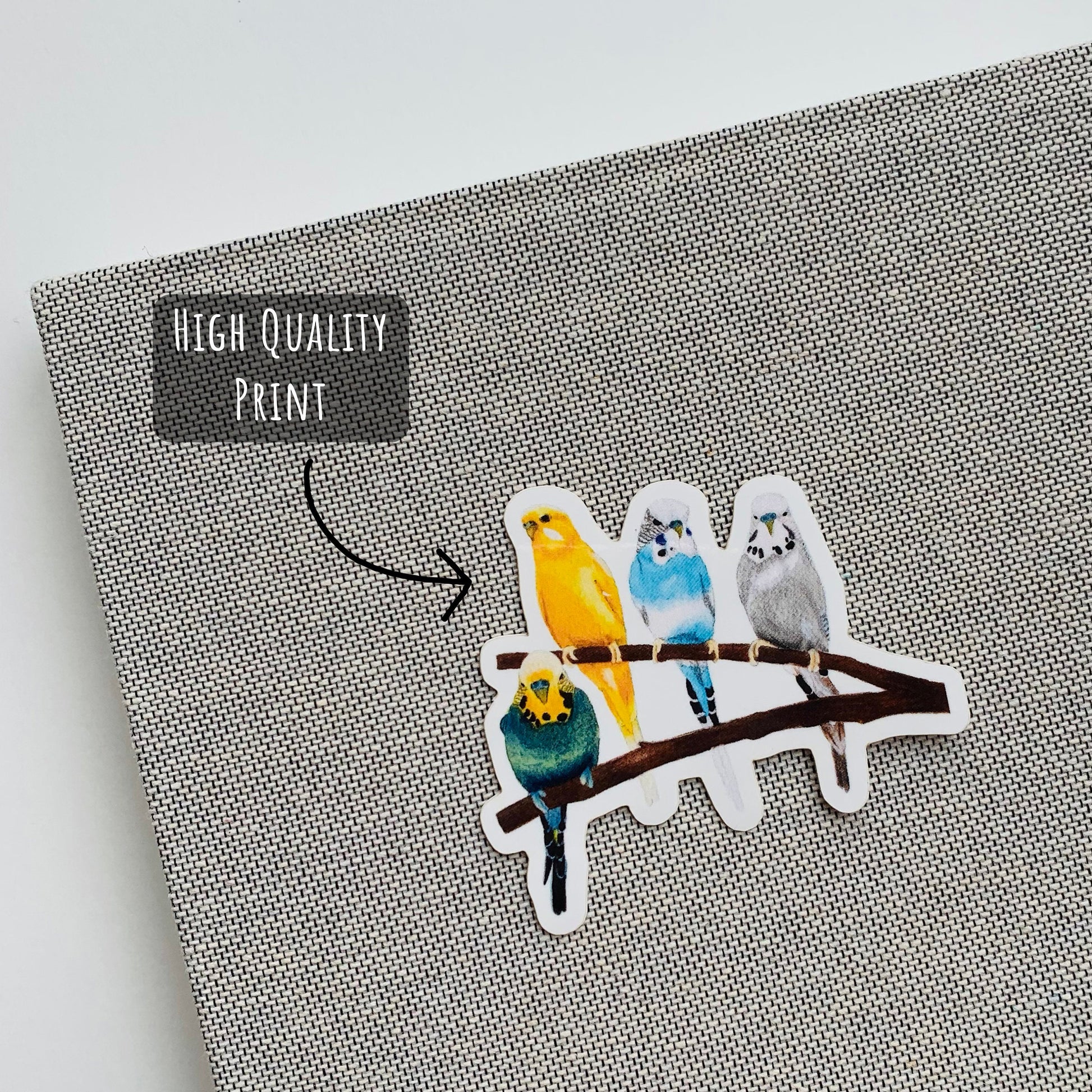 Vibrant Budgie Birds sticker placed on the front of a grey notebook with the text high quality print.