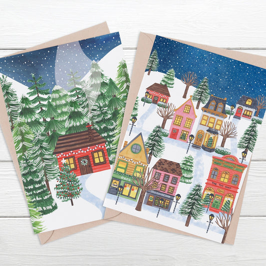 Two illustrated Christmas cards featuring a bright red log cabin with Christmas tree in front and snow sky, and a snowy colorful village scene with trees and a snow sky. Each card has a kraft paper envelope.