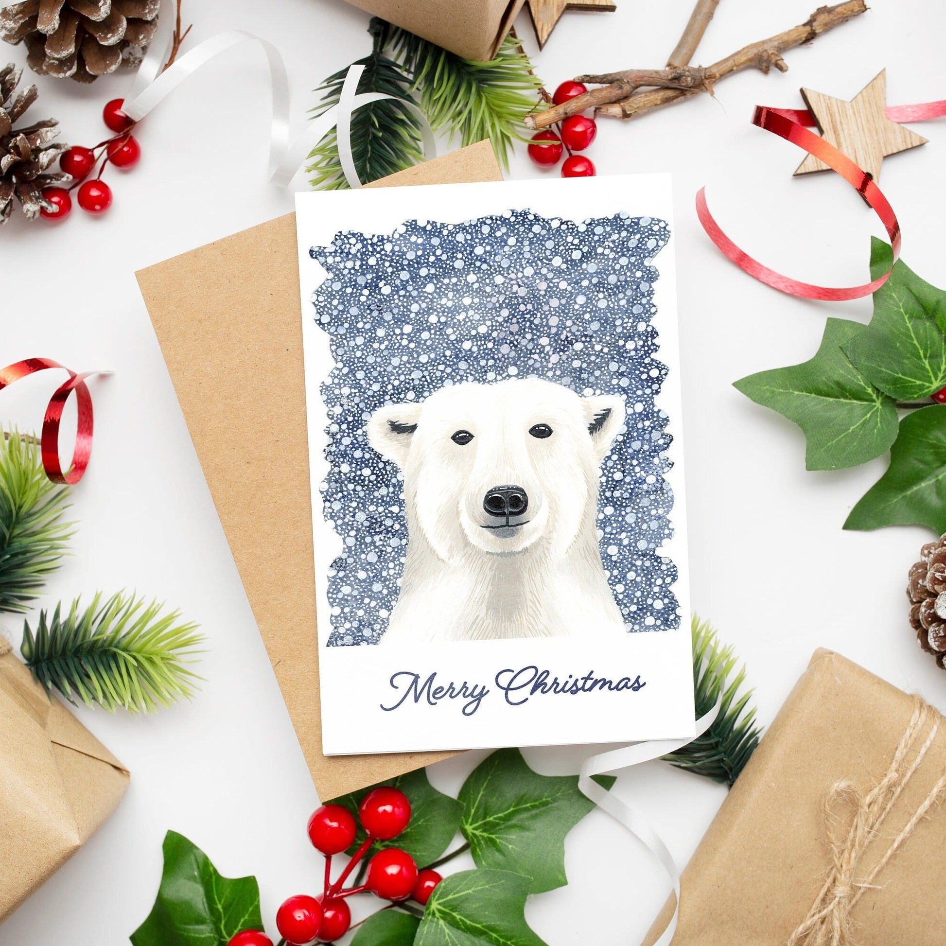 A Christmas card featuring a white polar bear on a dark blue snowy background with 'Merry Christmas' text at the bottom. The card is accompanied by festive decorations and comes with a kraft envelope.