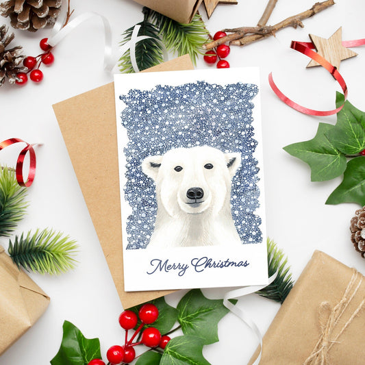 A Christmas card featuring a white polar bear on a dark blue snowy background with 'Merry Christmas' text at the bottom. The card is accompanied by festive decorations and comes with a kraft envelope.