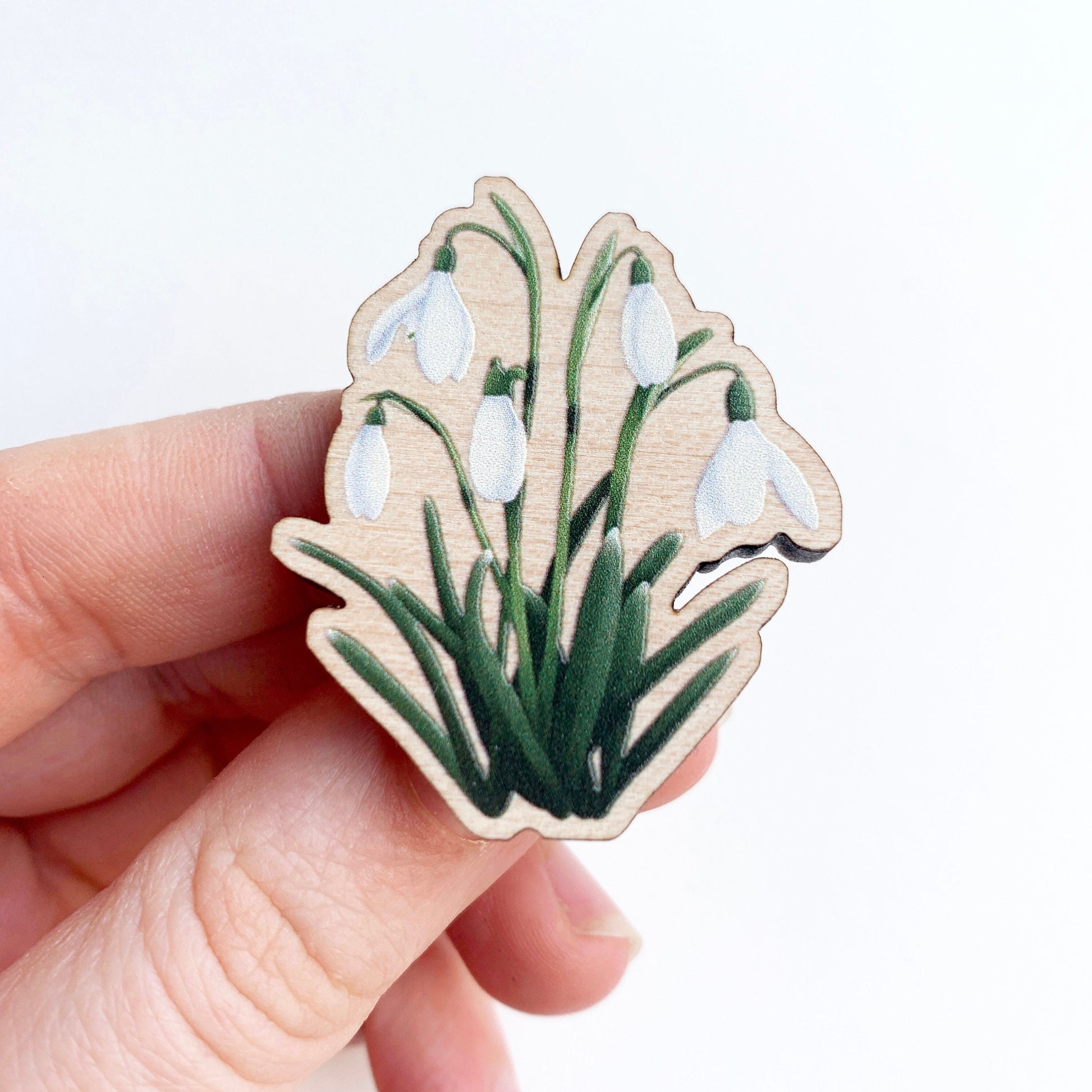 A hand holding a wooden pin badge with an illustration of white snowdrop flowers with stems and leaves. with a white background.