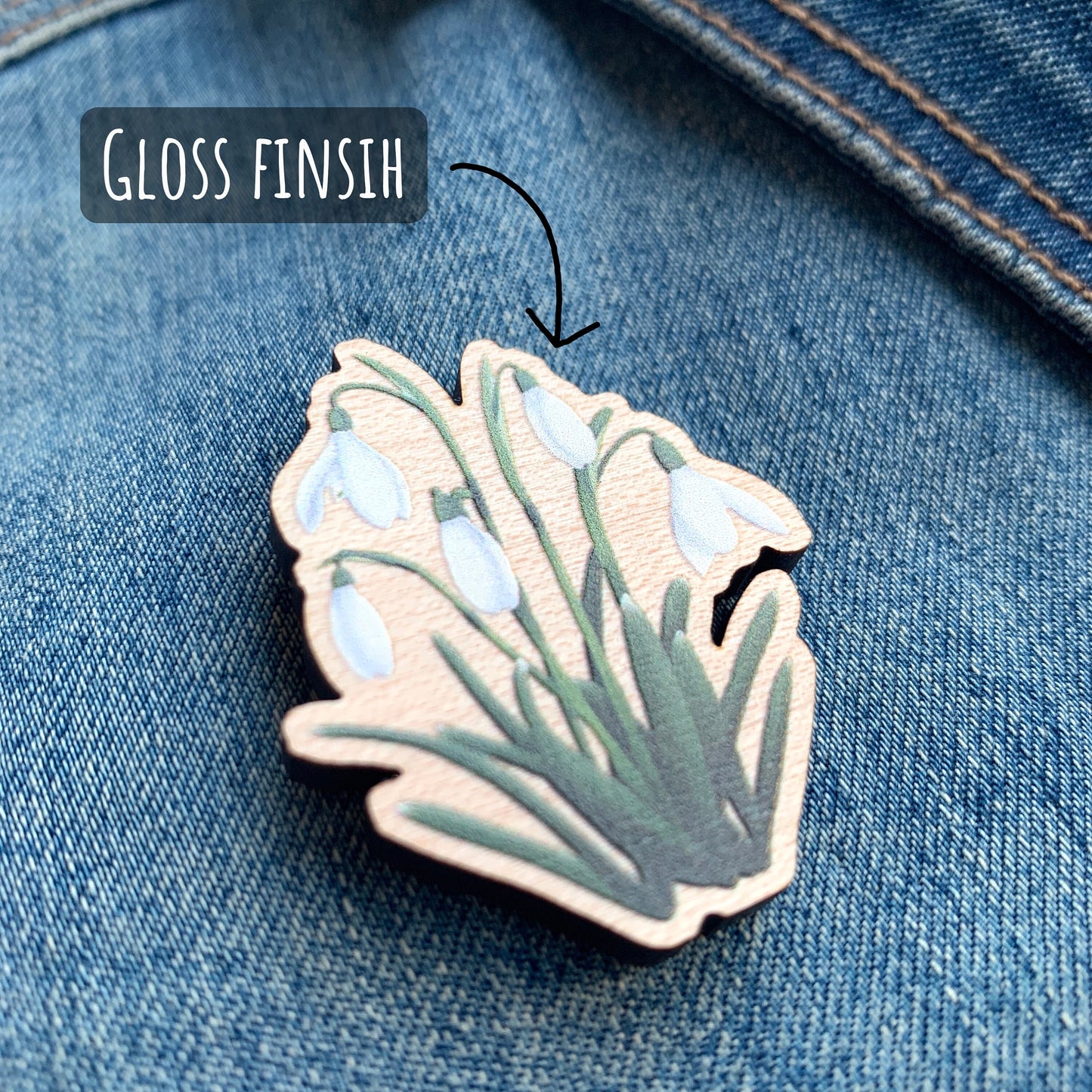 Wildflower Snowdrop Wooden Pin Badge close up showing the details and finish of the illustration and pin badge. Pin badge is placed on a blue denim jacket with the text gloss finish.