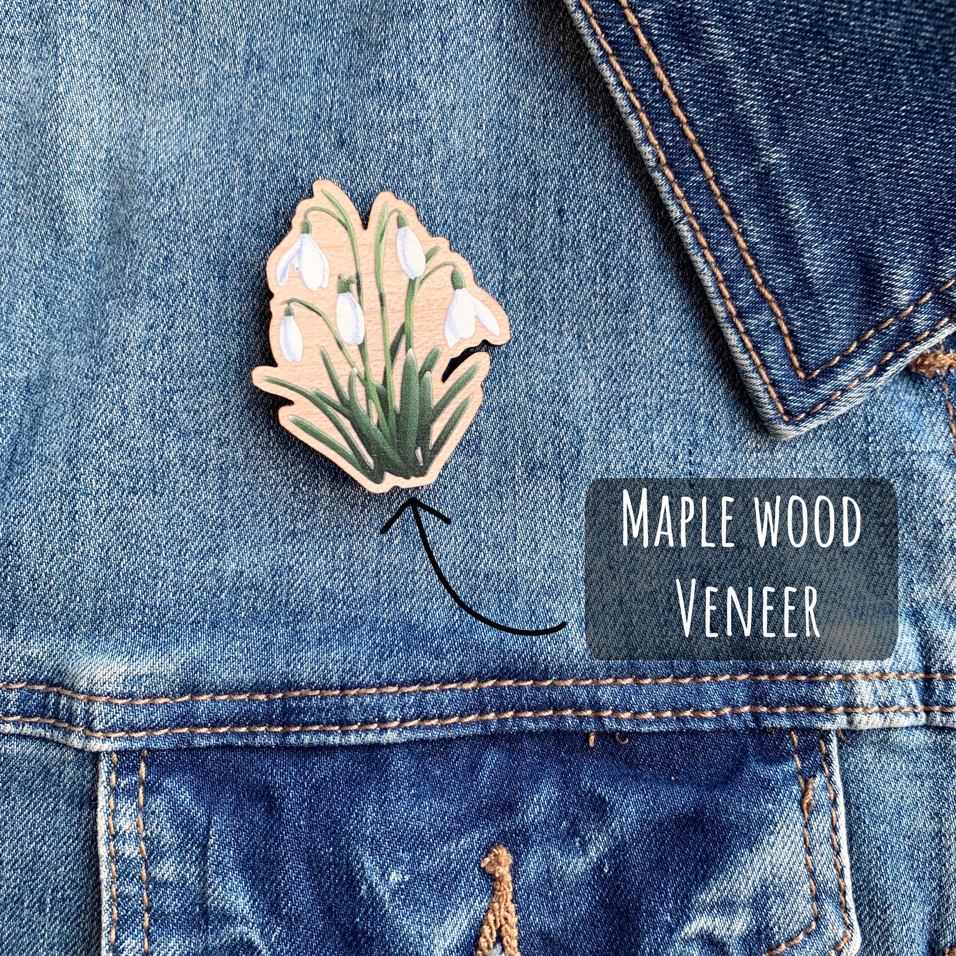 Wildflower Snowdrop Wooden Pin Badge placed on to a blue denim jacket with the text maple wood veneer.