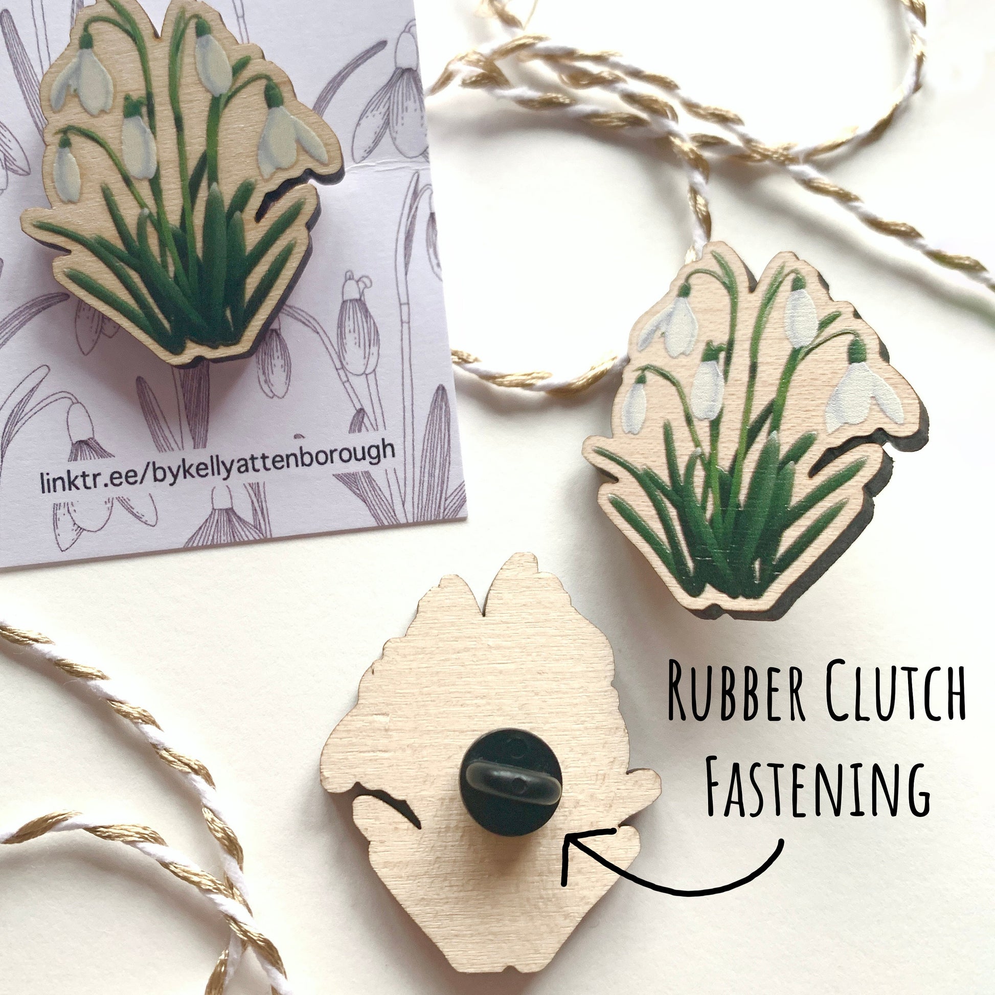 showing three Wildflower Snowdrop Wooden Pin Badges. One on its backing card one without and the third showing the back with the balck rubber clutch fastening. all on a white surface with the text rubber clutch fastening.