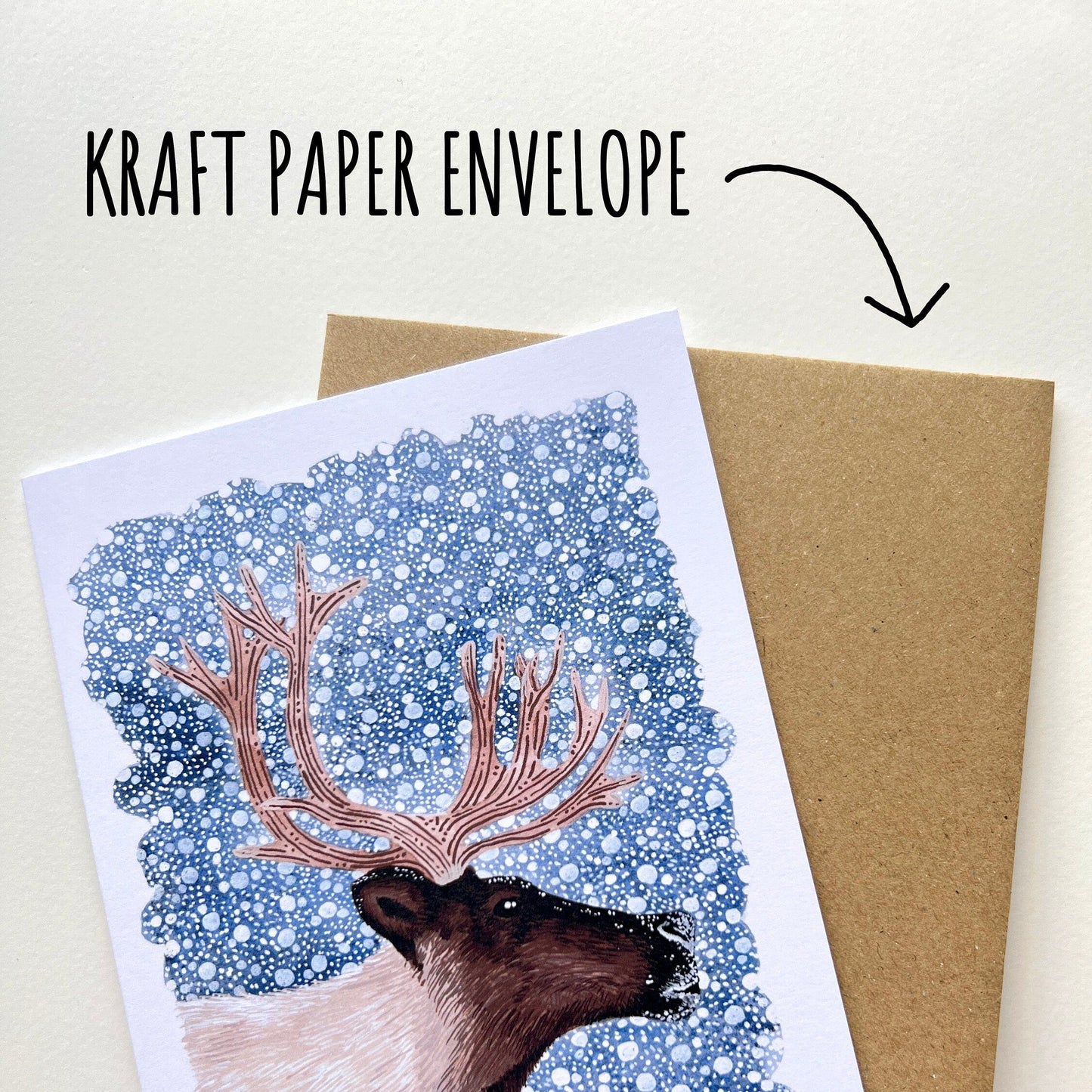 Greeting card with reindeer illustration and 'Merry Christmas' text, held over a kraft paper envelope. On a white background