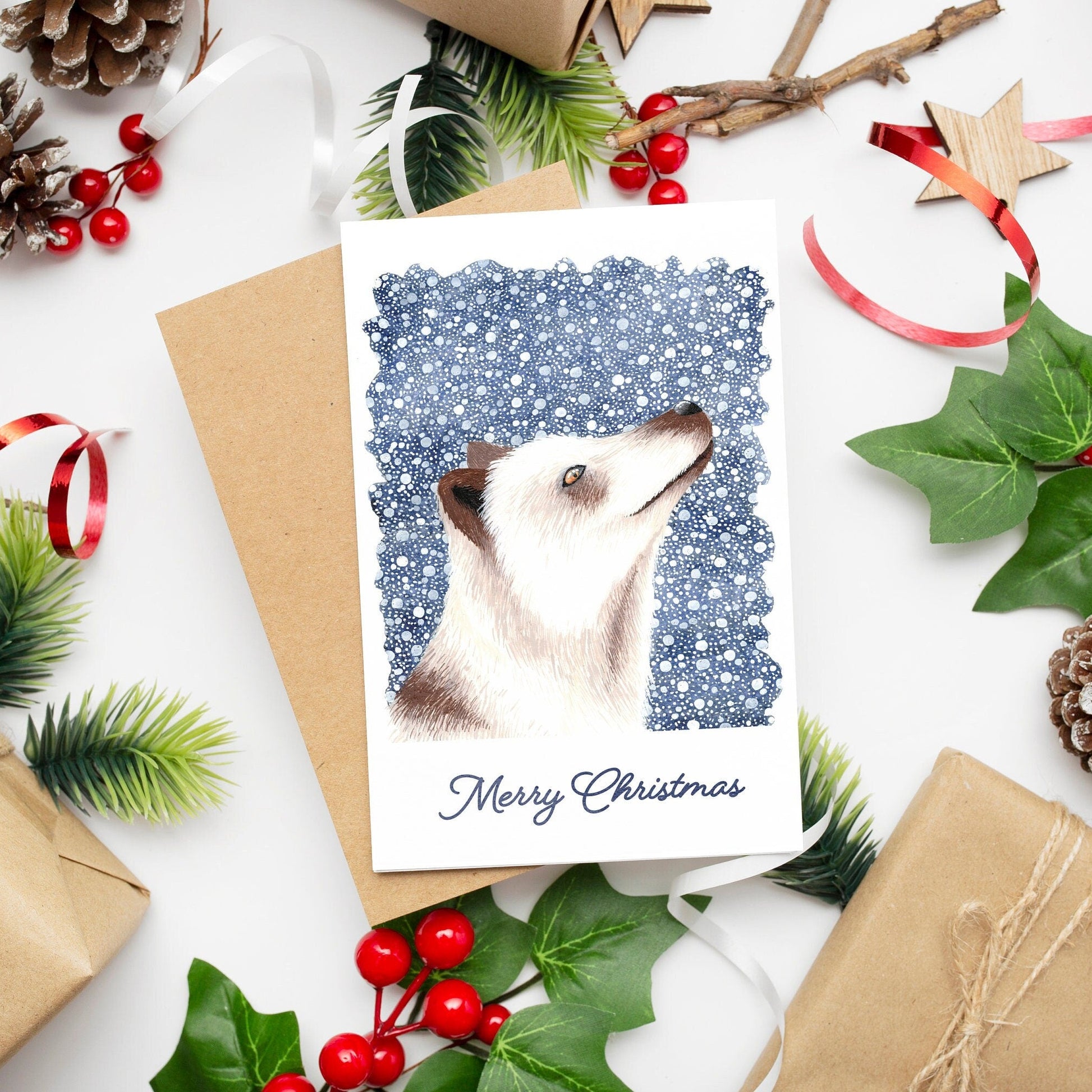 A Christmas card featuring a white arctic fox on a dark blue snowy background with 'Merry Christmas' text, presented on a table with festive decorations.
