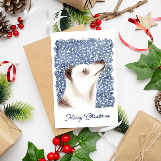 A Christmas card featuring a white arctic fox on a dark blue snowy background with 'Merry Christmas' text, presented on a table with festive decorations.