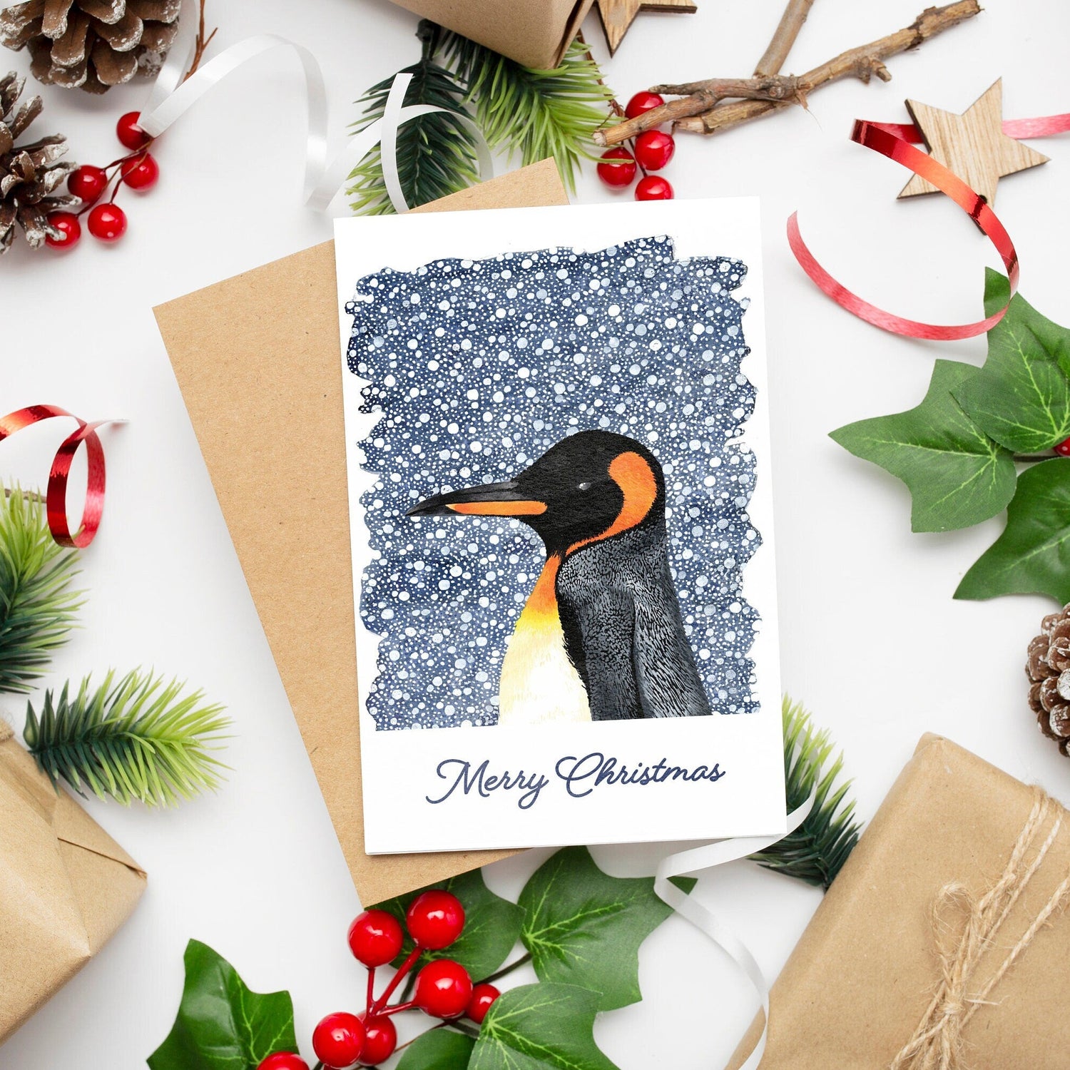 A Christmas card featuring an illustration of a penguin on a dark blue snowy background with 'Merry Christmas' text, surrounded by festive decorations and a kraft envelope.