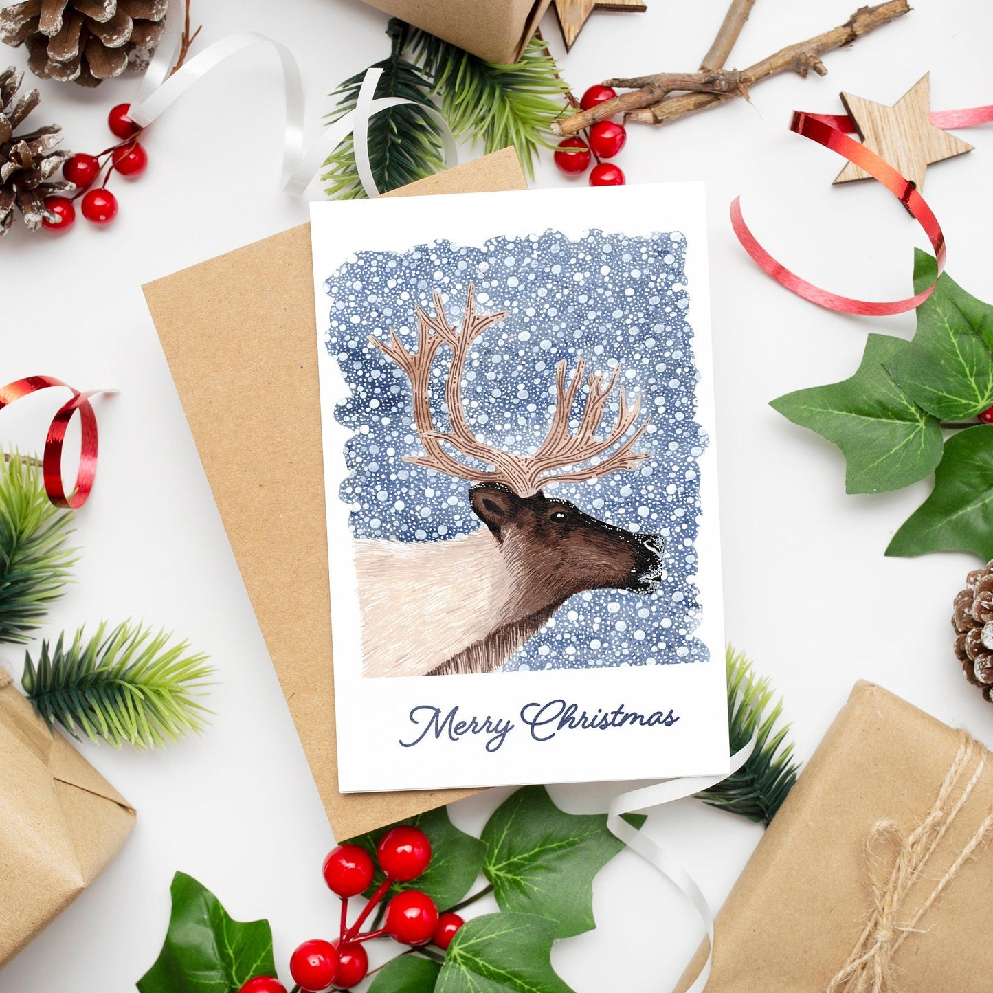 A Christmas card featuring a reindeer on a dark blue snowy background with 'Merry Christmas' text, presented with festive decorations and a kraft envelope.