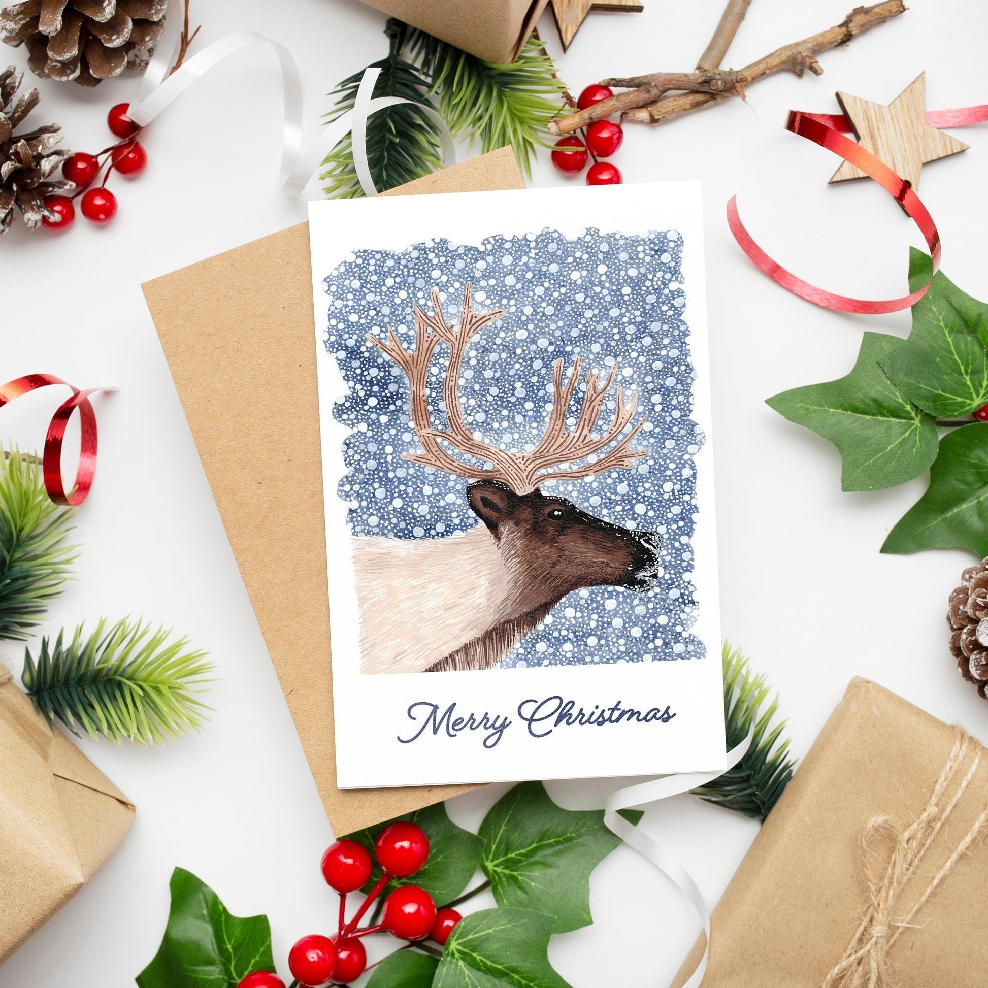 A Christmas card featuring a reindeer on a dark blue snowy background with 'Merry Christmas' text, presented with festive decorations and a kraft envelope.