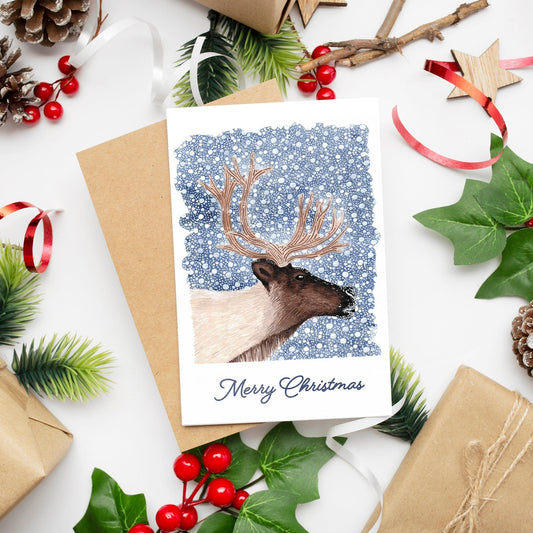 A Christmas card featuring a reindeer on a dark blue snowy background with 'Merry Christmas' text, presented with festive decorations and a kraft envelope.