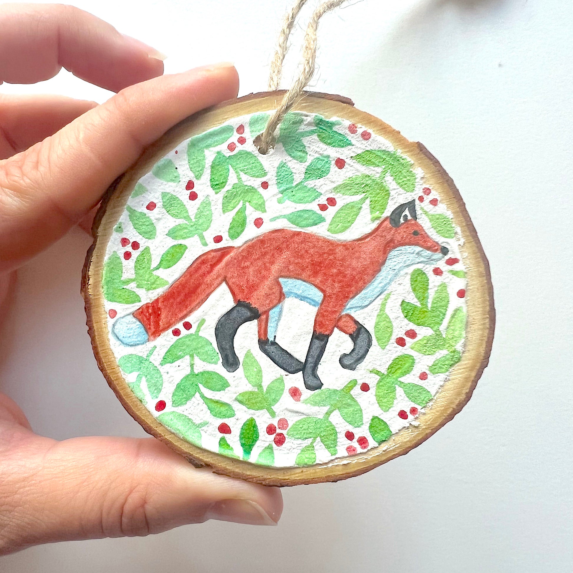 Hand-painted wooden slice Christmas decorations featuring various woodland animal designs including a fox, bear, deer, robin, rabbit, and squirrel, surrounded by green and red painted foliage.