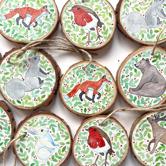 Hand-painted wooden slice Christmas decorations featuring various woodland animal designs including a fox, bear, deer, robin, rabbit, and squirrel, surrounded by green and red painted foliage.