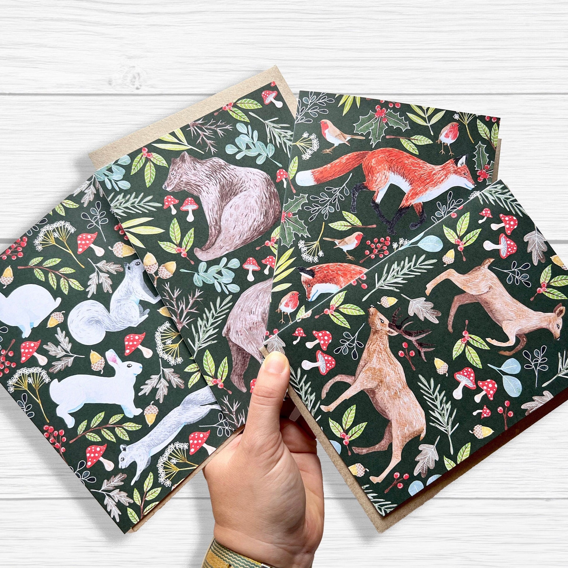 A set of Woodland Animal Christmas cards  featuring foxes, deer, bears, and squirrel with rabbits. held by a hand with a white wood backgorund.