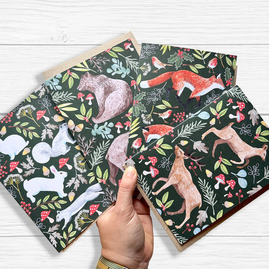 A set of Woodland Animal Christmas cards  featuring foxes, deer, bears, and squirrel with rabbits. held by a hand with a white wood backgorund.