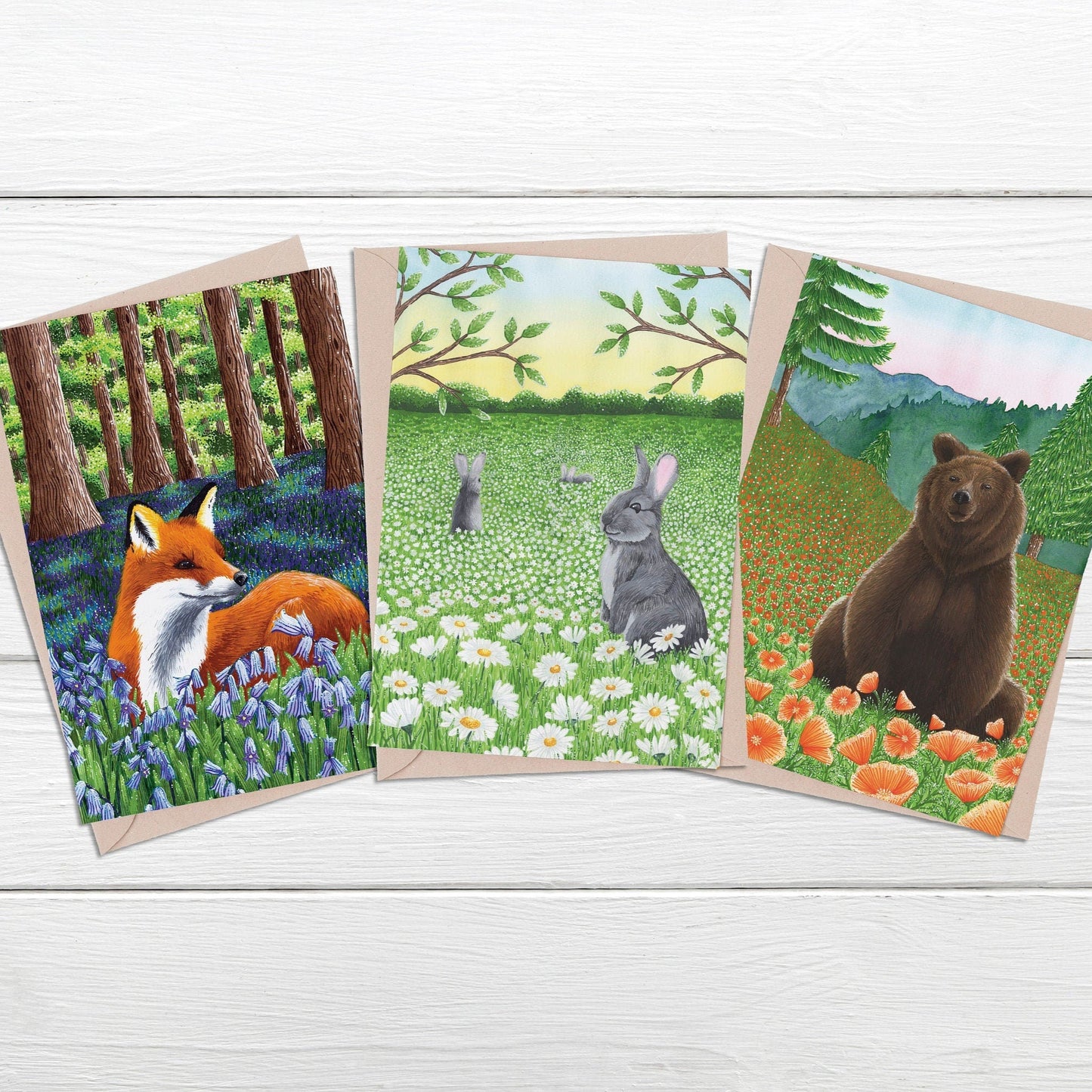 Three greeting cards featuring a fox, rabbit, and bear in natural settings on a wooden surface.