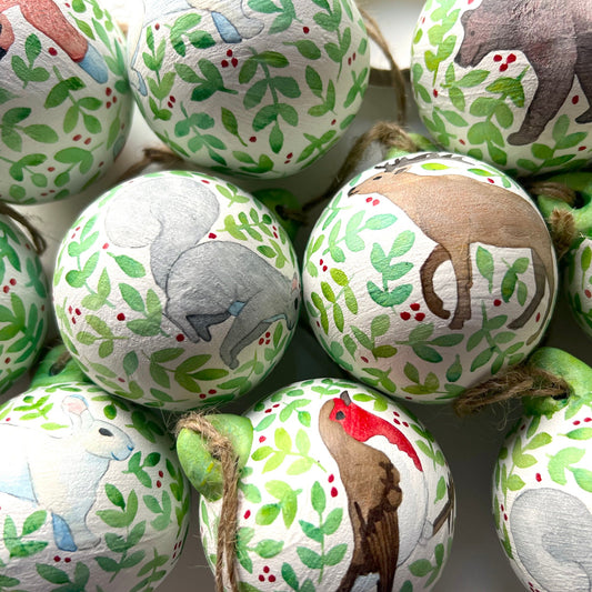 A collection of hand-painted ceramic Christmas baubles with various woodland animal designs including a fox, bear, deer, robin, rabbit, and squirrel, surrounded by green blue leaves and red berries.