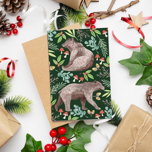A Christmas card featuring an illustration of two bears surrounded by festive foliage, presented on a table with holiday decorations.