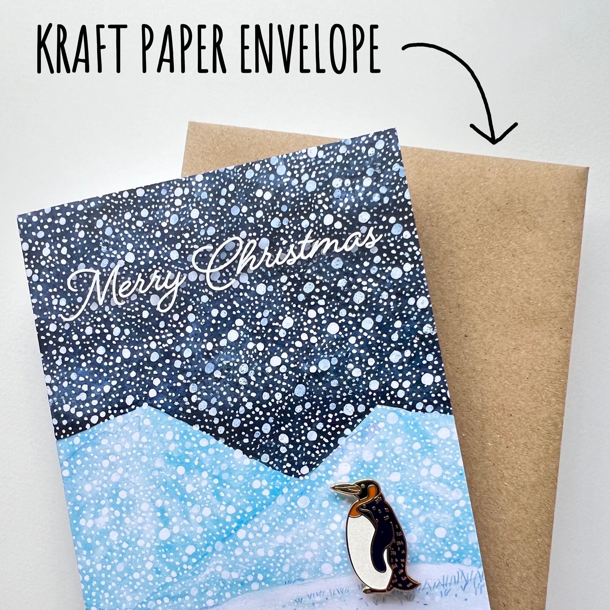 A snowy mountain Christmas card with a penguin  penguin pin badge and a kraft paper envelope on a white background with the font Kraft paper envelope.