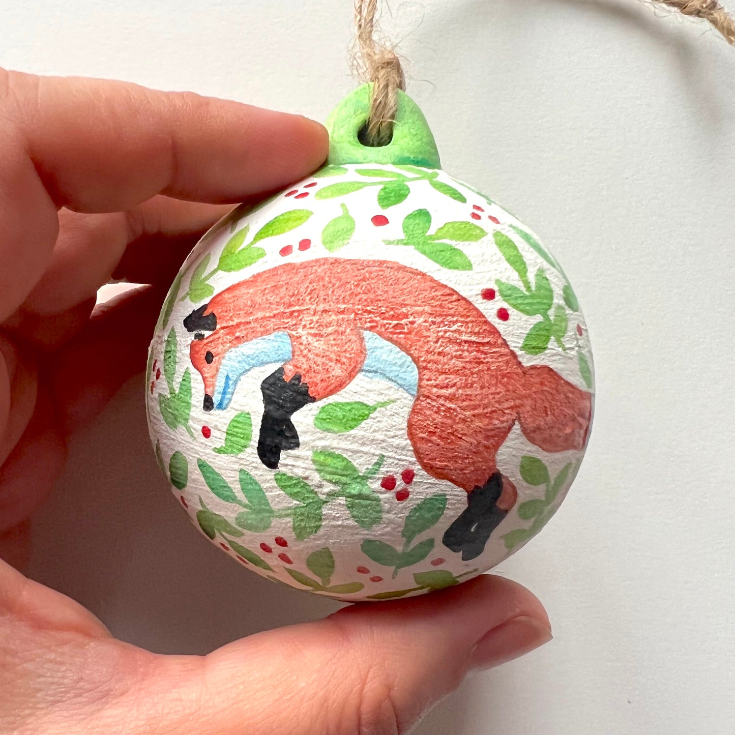 A collection of hand-painted ceramic Christmas baubles with various woodland animal designs including a fox, bear, deer, robin, rabbit, and squirrel, surrounded by green blue leaves and red berries.